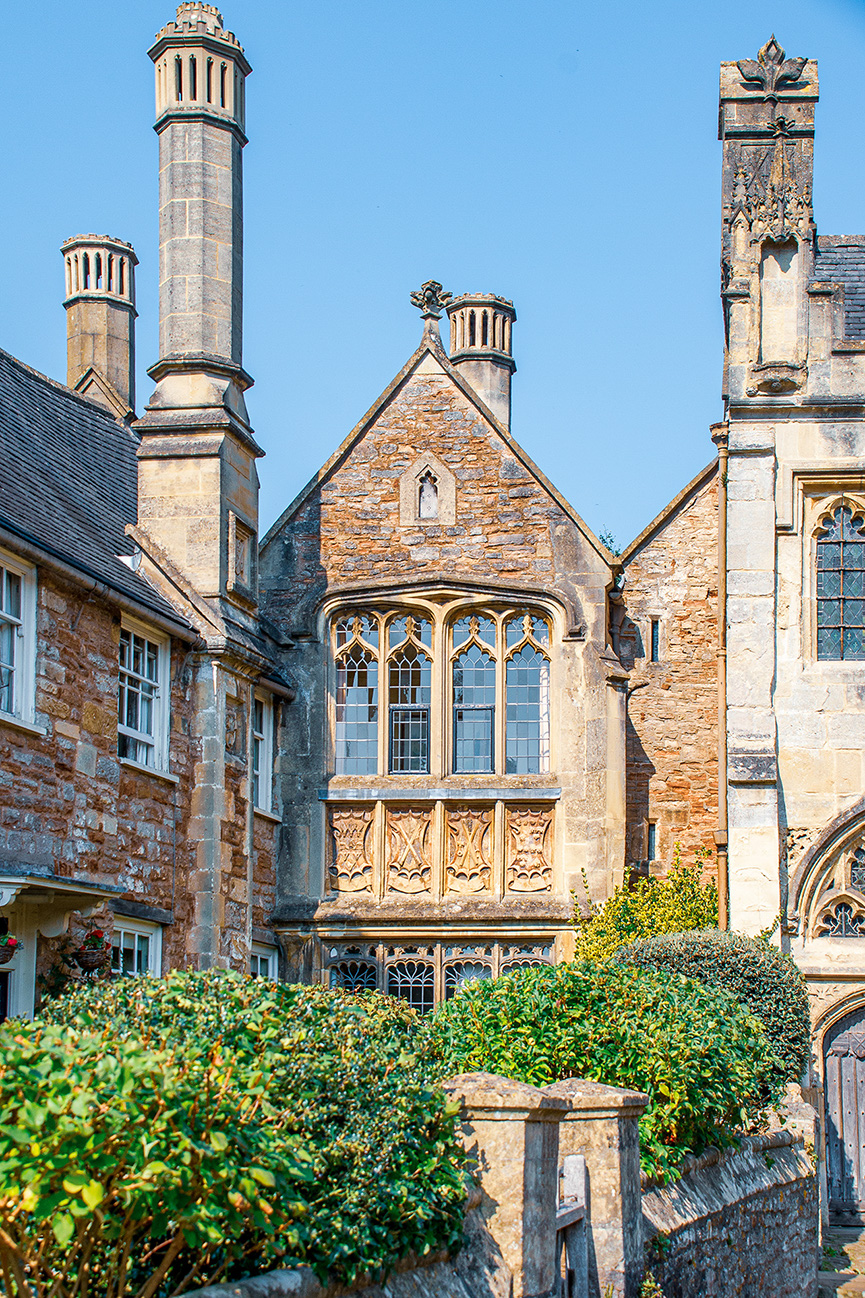 The image depicts a historic building with intricate stone architecture, featuring tall chimneys, ornate windows, and detailed stone carvings. The building is surrounded by lush greenery and appears to be part of a larger complex, possibly a university or a historic site..