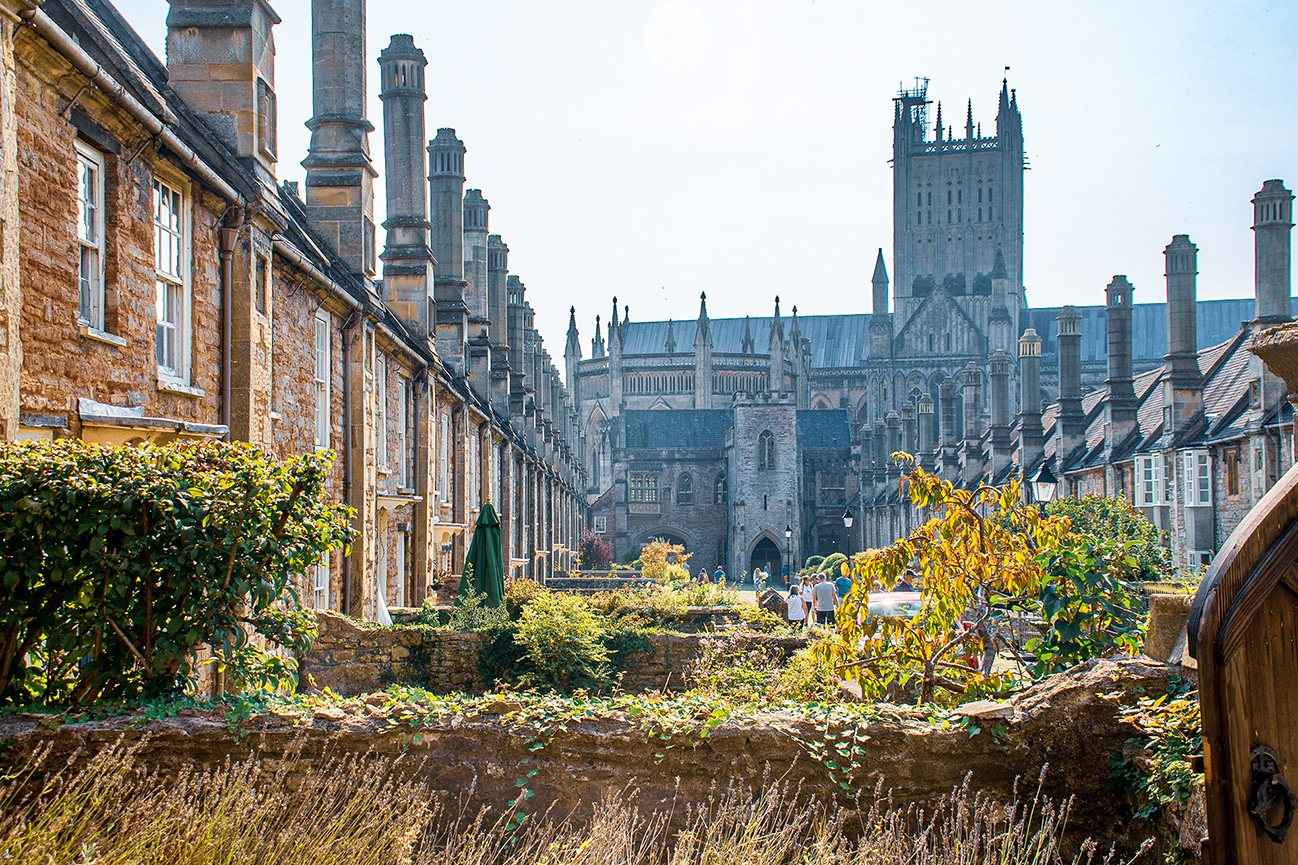The image depicts a scenic view of a historic university campus, featuring traditional stone buildings with Gothic architecture. The central focus is a large, ornate cathedral or chapel with intricate details and towering spires. The foreground includes lush greenery and a pathway where people are walking, adding a sense of scale and liveliness to the scene. The overall atmosphere is serene and scholarly, characteristic of an old, prestigious academic institution.
