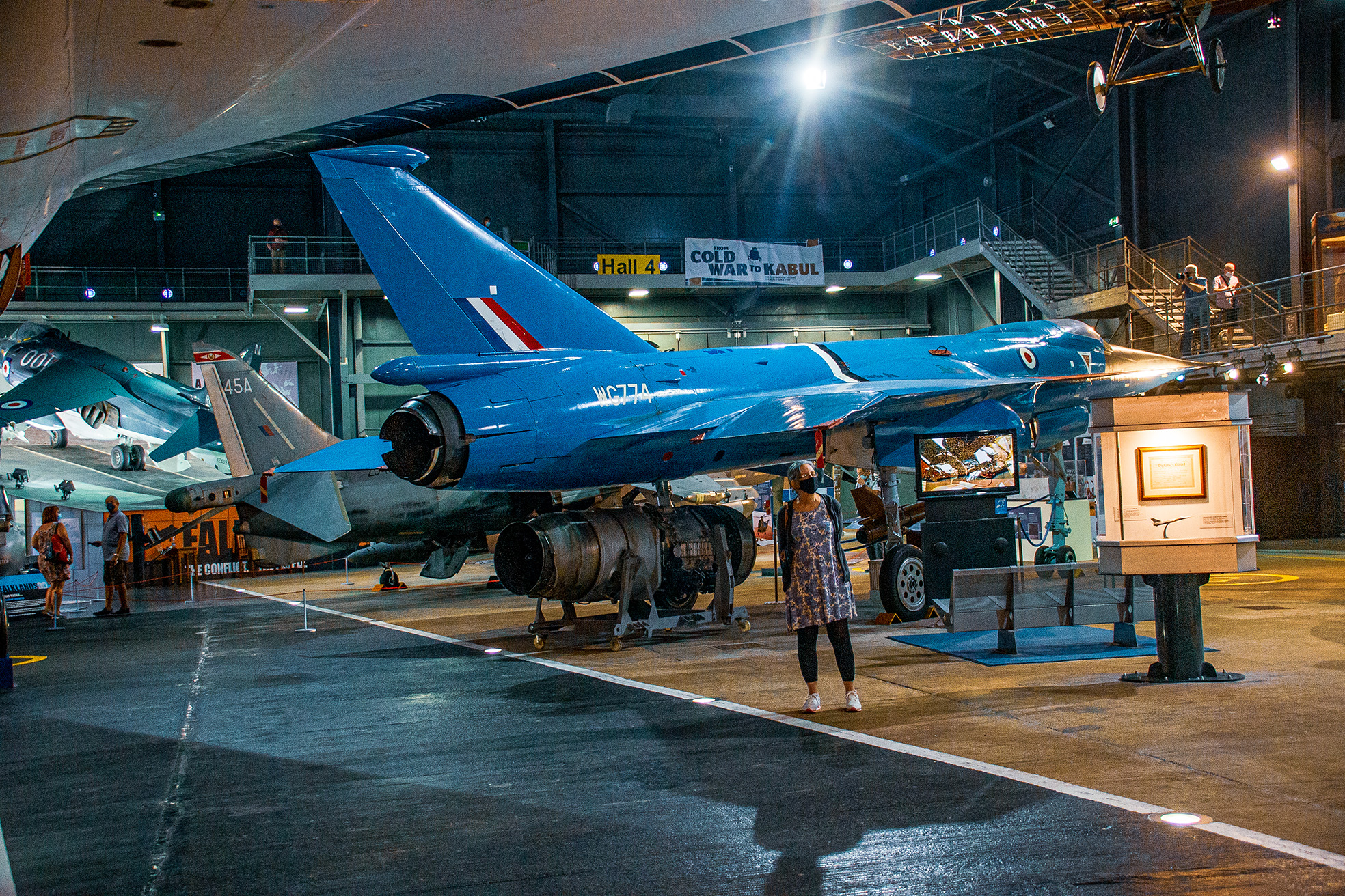 The image depicts an indoor aviation exhibit featuring a blue fighter jet on display. The jet is mounted on a stand with informational displays around it. Several people are present, observing the exhibit. The setting appears to be a large hangar or museum space, with high ceilings and industrial lighting. A sign indicating 'Hall 4' and a banner reading 'From Cold War to Kabul' are visible in the background.
