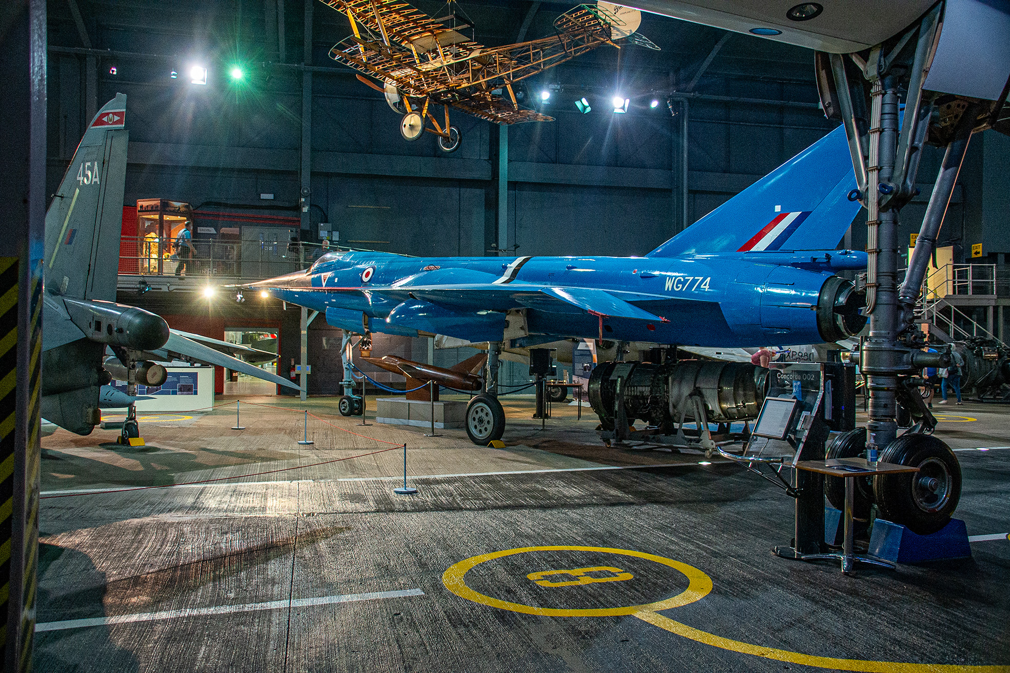 The image depicts an aircraft hangar with a prominent blue jet, marked with WG774, on display. The jet is elevated on supports, showcasing its undercarriage. In the background, there is a vintage biplane suspended from the ceiling. The hangar also features various other aircraft components and exhibits, with informational placards visible near the blue jet. The setting is well-lit, highlighting the aircraft and creating a museum-like atmosphere.