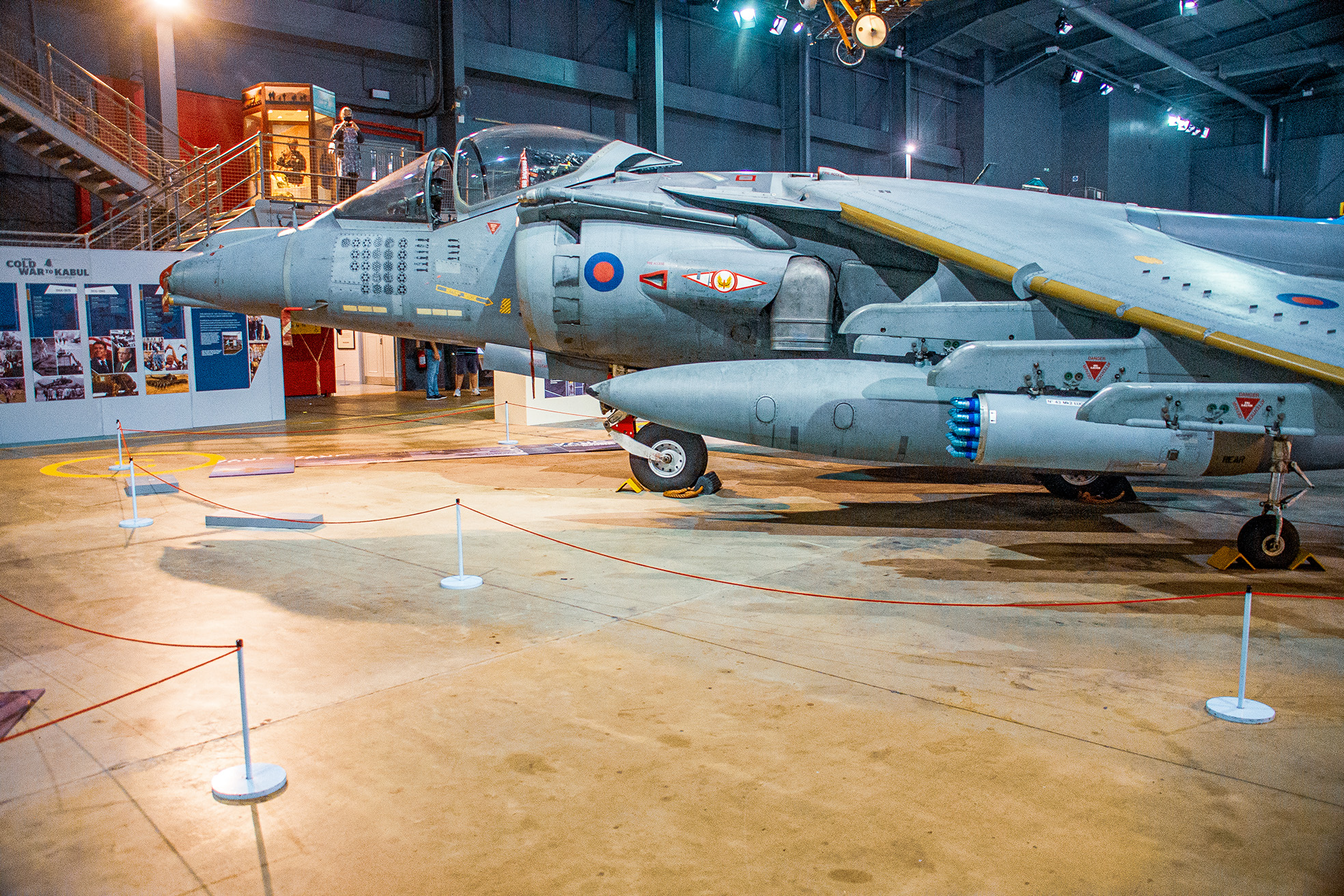 The image depicts a military aircraft on display in a museum. The aircraft is a SEPECAT Jaguar, identifiable by its distinctive shape and markings. It is equipped with various weapons and missiles, showcasing its role as a ground attack aircraft. The display is cordoned off with red ropes and white posts to prevent visitors from touching the exhibit. The background shows informational panels and a staircase leading to an upper viewing area.