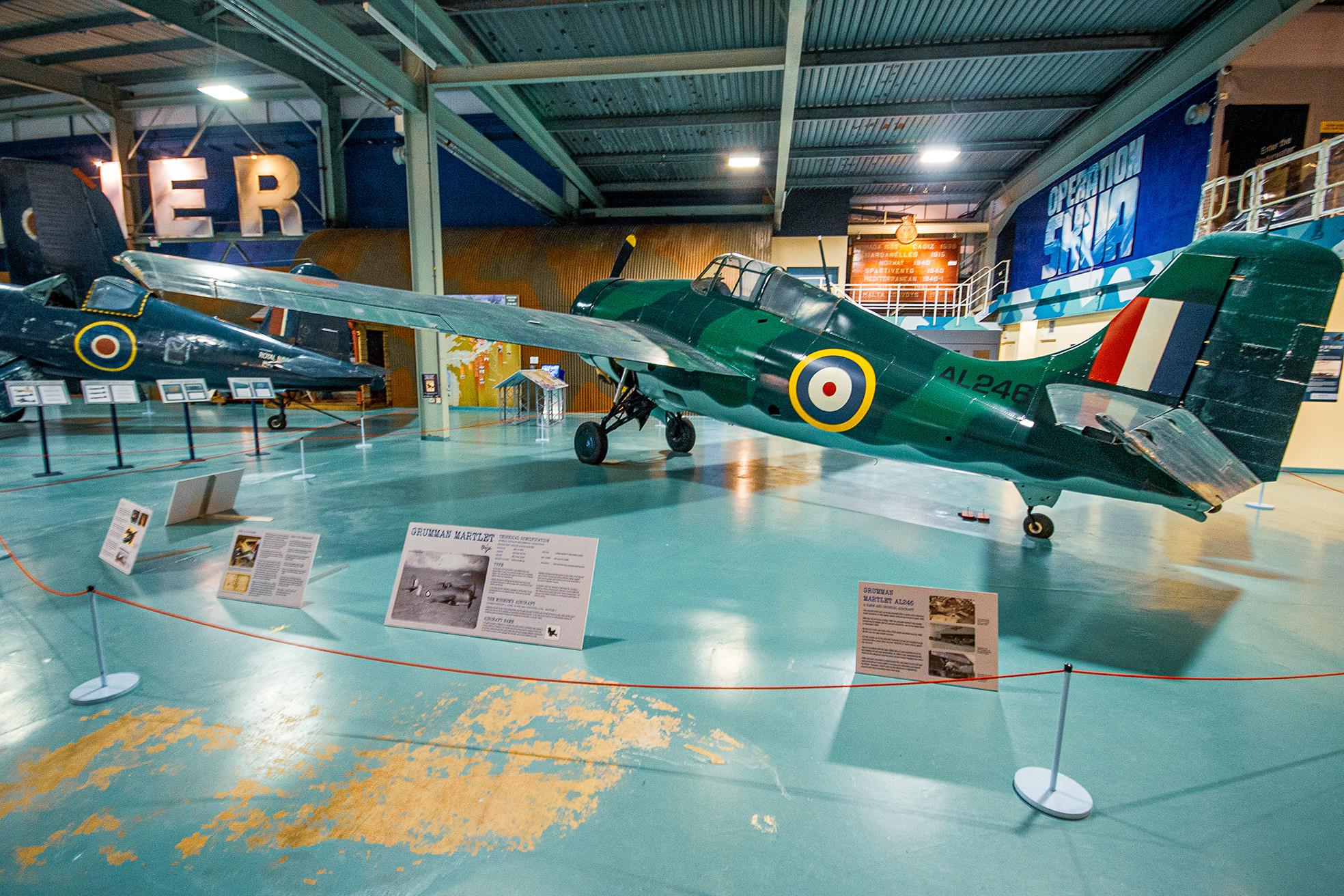 The image depicts a museum exhibit featuring a Grumman Martlet, a World War II aircraft. The aircraft is displayed indoors with informational placards around it, providing details about its history and specifications. The plane is painted in a green color scheme with distinctive roundels on its wings and fuselage. The museum setting includes other aircraft and exhibits in the background, with a high ceiling and large windows.