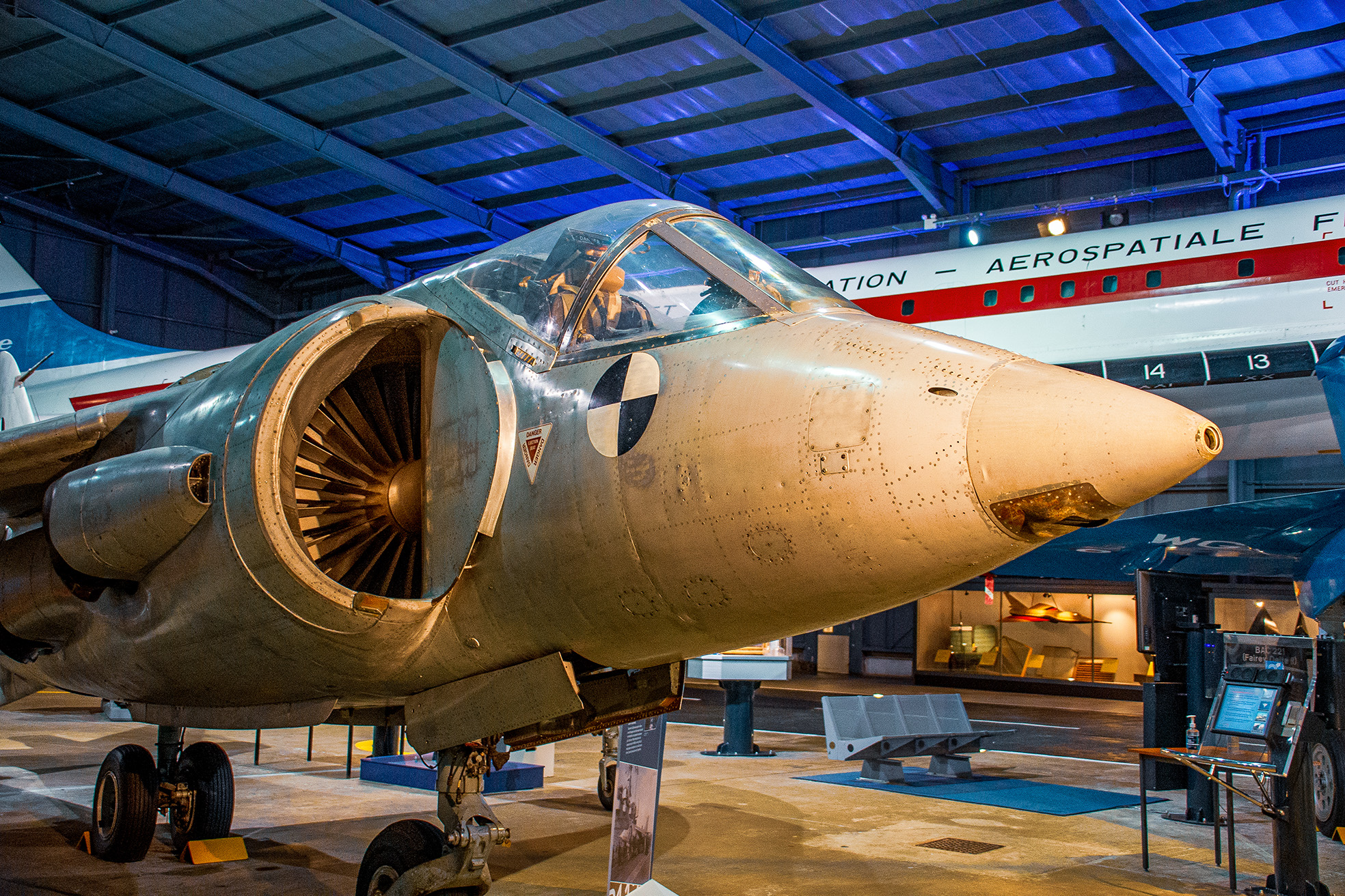 The image shows a military aircraft on display in a museum. The aircraft has a single large engine with a prominent circular intake and a jet exhaust nozzle. The nose of the aircraft is painted with a distinctive roundel marking. The display area includes informational placards and is well-lit, with other aircraft visible in the background. The setting appears to be an aviation museum, showcasing historical aircraft