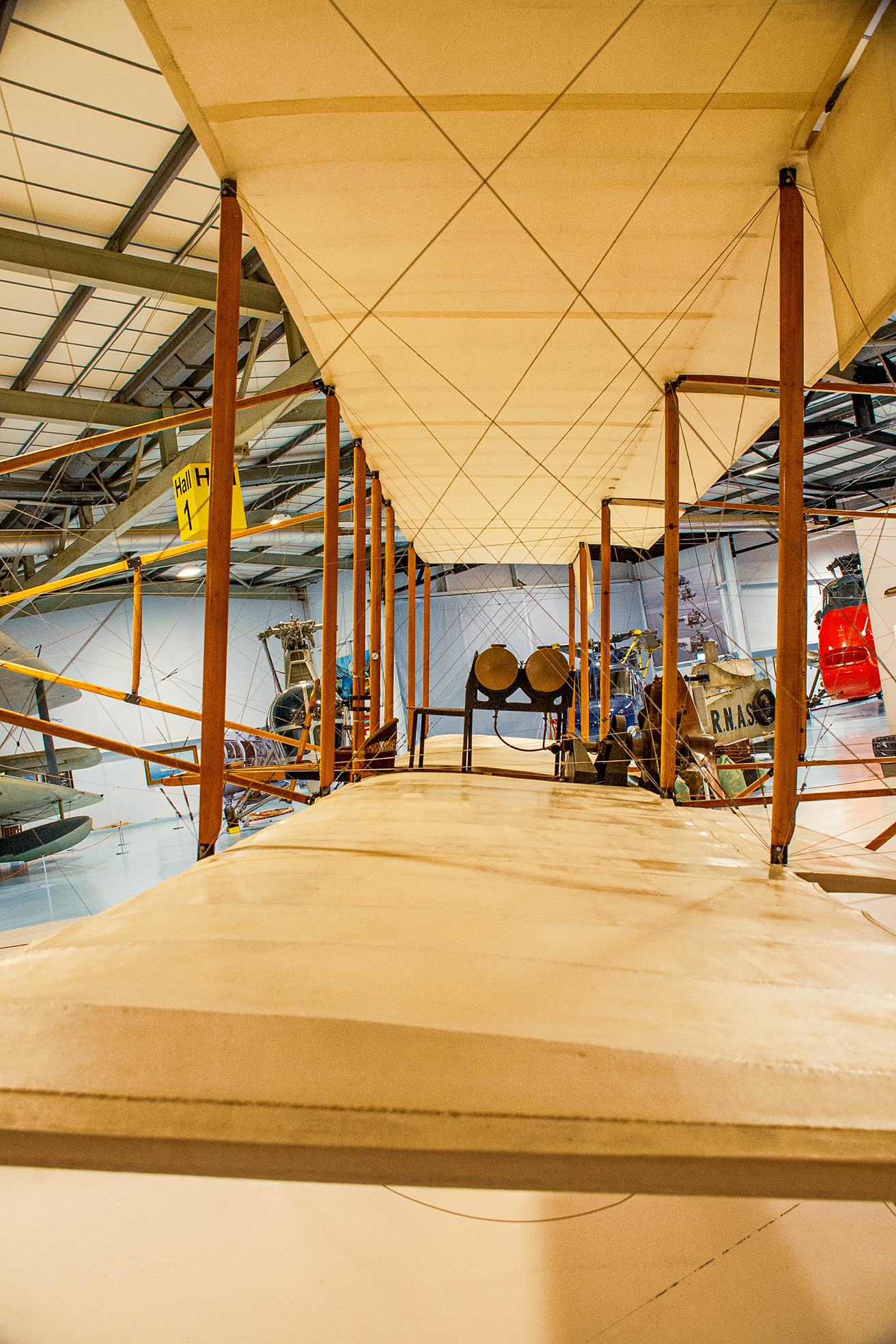 The image depicts an interior view of an aviation museum, showcasing an early aircraft suspended from the ceiling. The aircraft has a wooden frame and fabric covering, indicative of early 20th-century design. The perspective is from the end of the wing of the aircraft, looking down its length towards the fuselage.