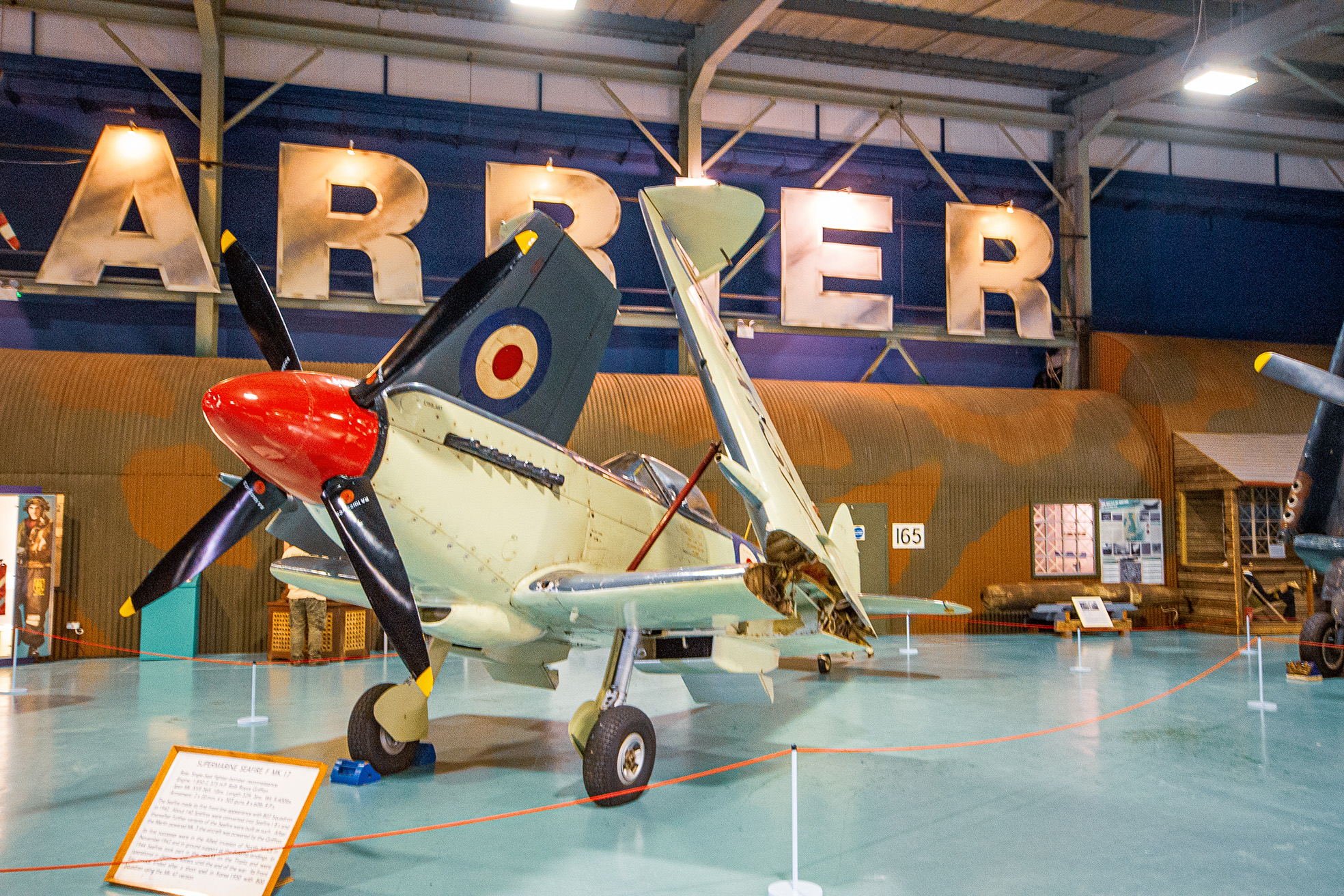 The image shows a vintage military aircraft displayed in a museum. The aircraft is a Supermarine Spitfire, identifiable by its distinctive elliptical wings and the roundel markings on its fuselage. The setting appears to be an indoor hangar with large letters spelling 'ABER' on the wall in the background. The aircraft is cordoned off with red ropes and has an information placard in front of it, suggesting it is part of an exhibit.