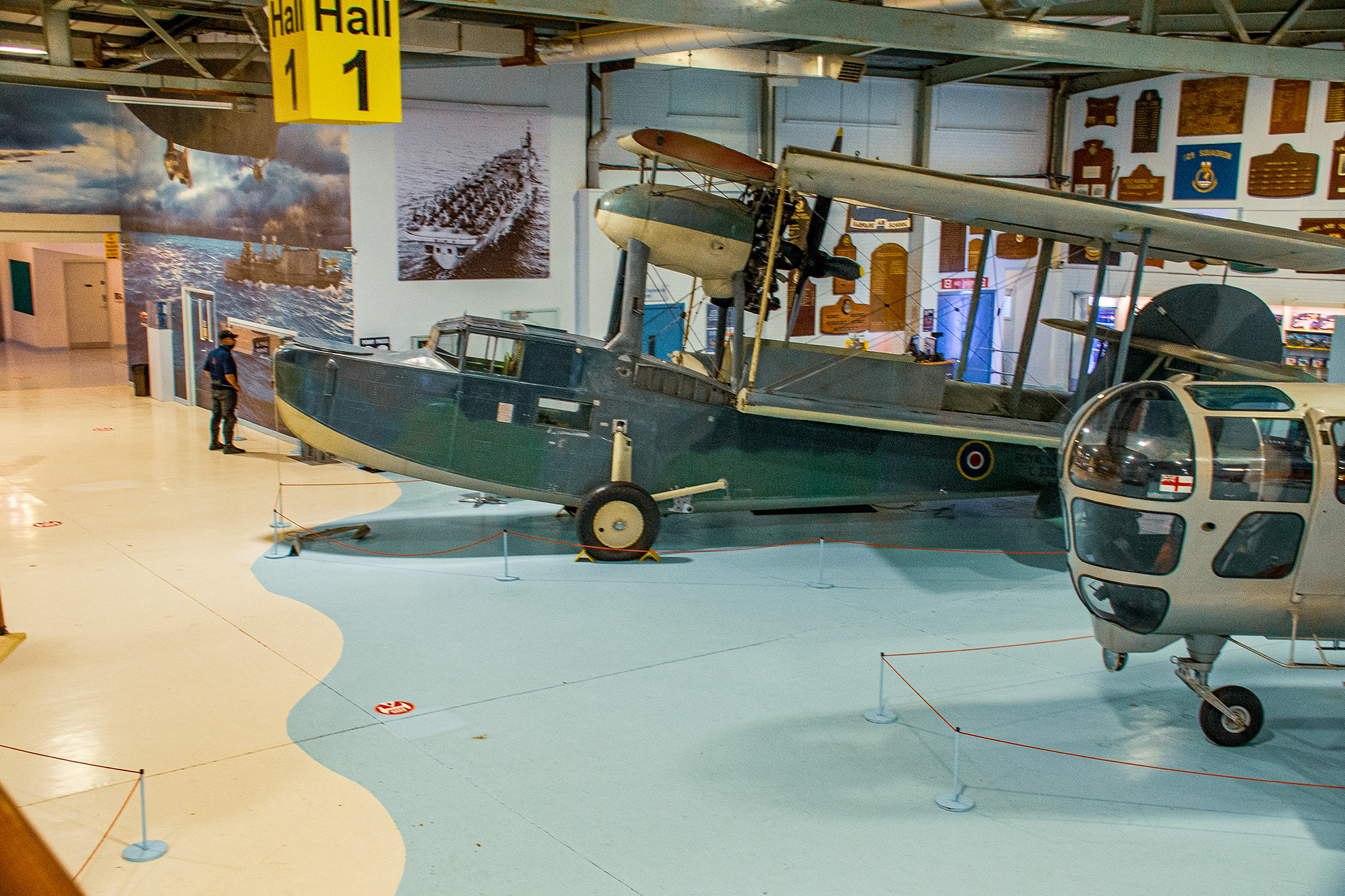 The image depicts an aviation museum exhibit featuring a vintage seaplane and a modern helicopter. The seaplane, with its distinctive floaters, is prominently displayed in the center, while the helicopter is positioned in the foreground. The museum hall is spacious with high ceilings, and various historical aviation artifacts and photographs adorn the walls. A sign indicating 'Hall 1' is visible, suggesting this is one of several exhibit halls in the museum. The overall atmosphere is one of historical preservation and educational display.