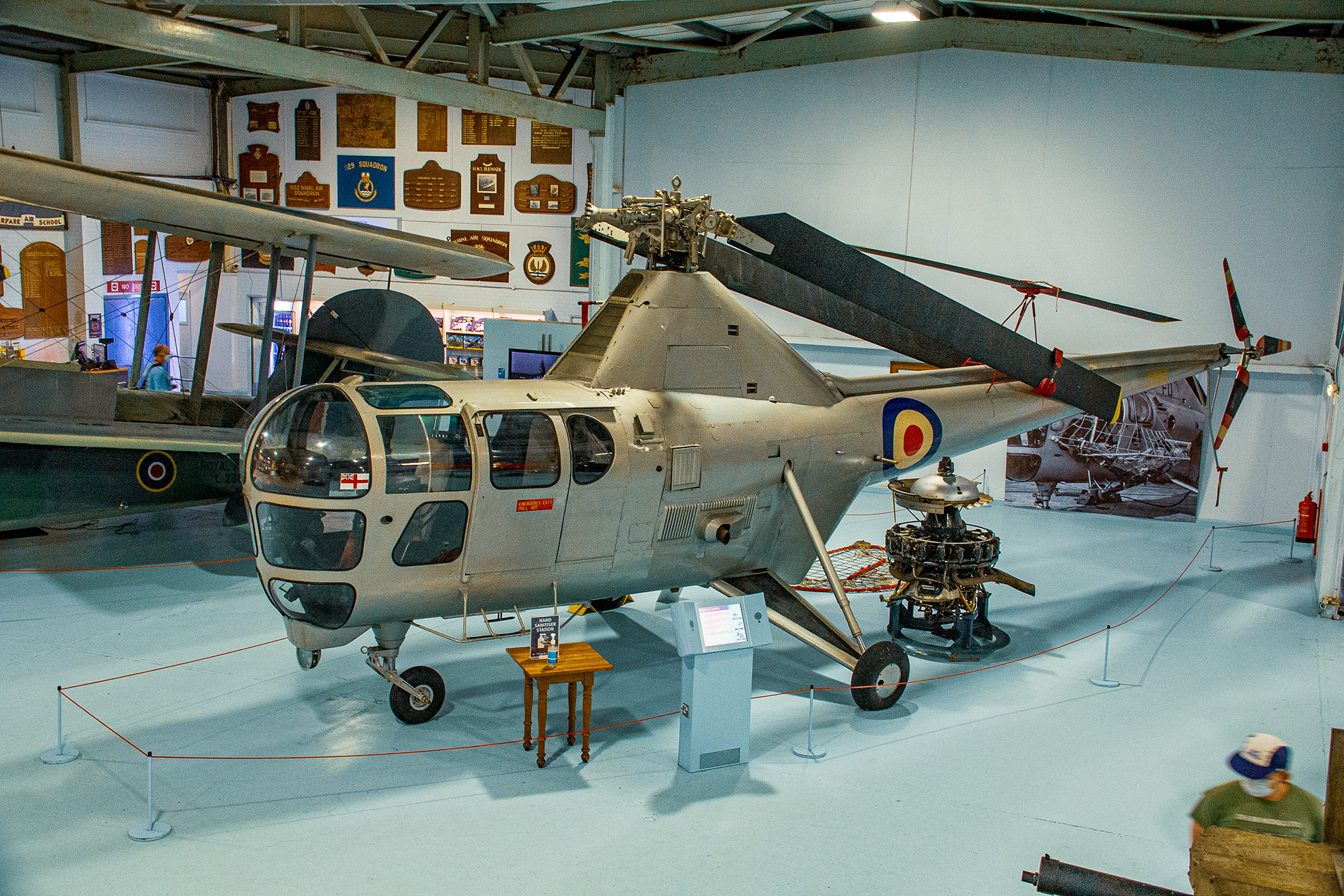 The image shows a vintage helicopter on display in a museum. The helicopter has a metallic finish with a roundel marking on its side, indicating it might be a military aircraft. It features a bubble cockpit, dual landing skids, and a visible engine mounted at the front. There is an informational display and a small table with a booklet next to the helicopter. The background includes various aviation artifacts and plaques.