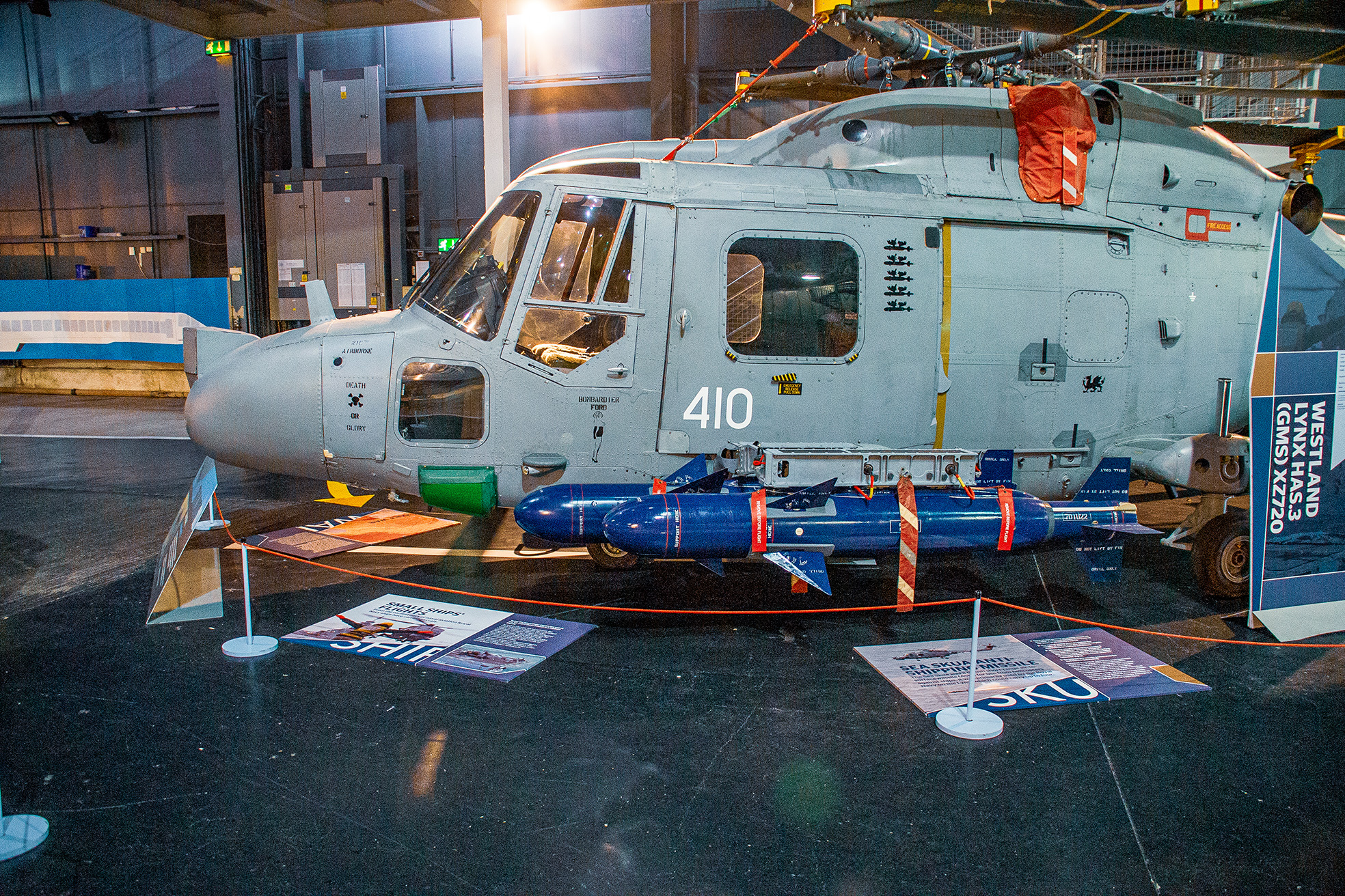 The image depicts a military helicopter on display in a museum or exhibition setting. The helicopter, marked with the number 410 and various insignia, is equipped with armaments attached to its underside. Informational placards and displays surround the helicopter, providing context and details about its history and specifications. The setting appears to be indoors, with the helicopter positioned on a polished floor and cordoned off with barriers and ropes to prevent close access.