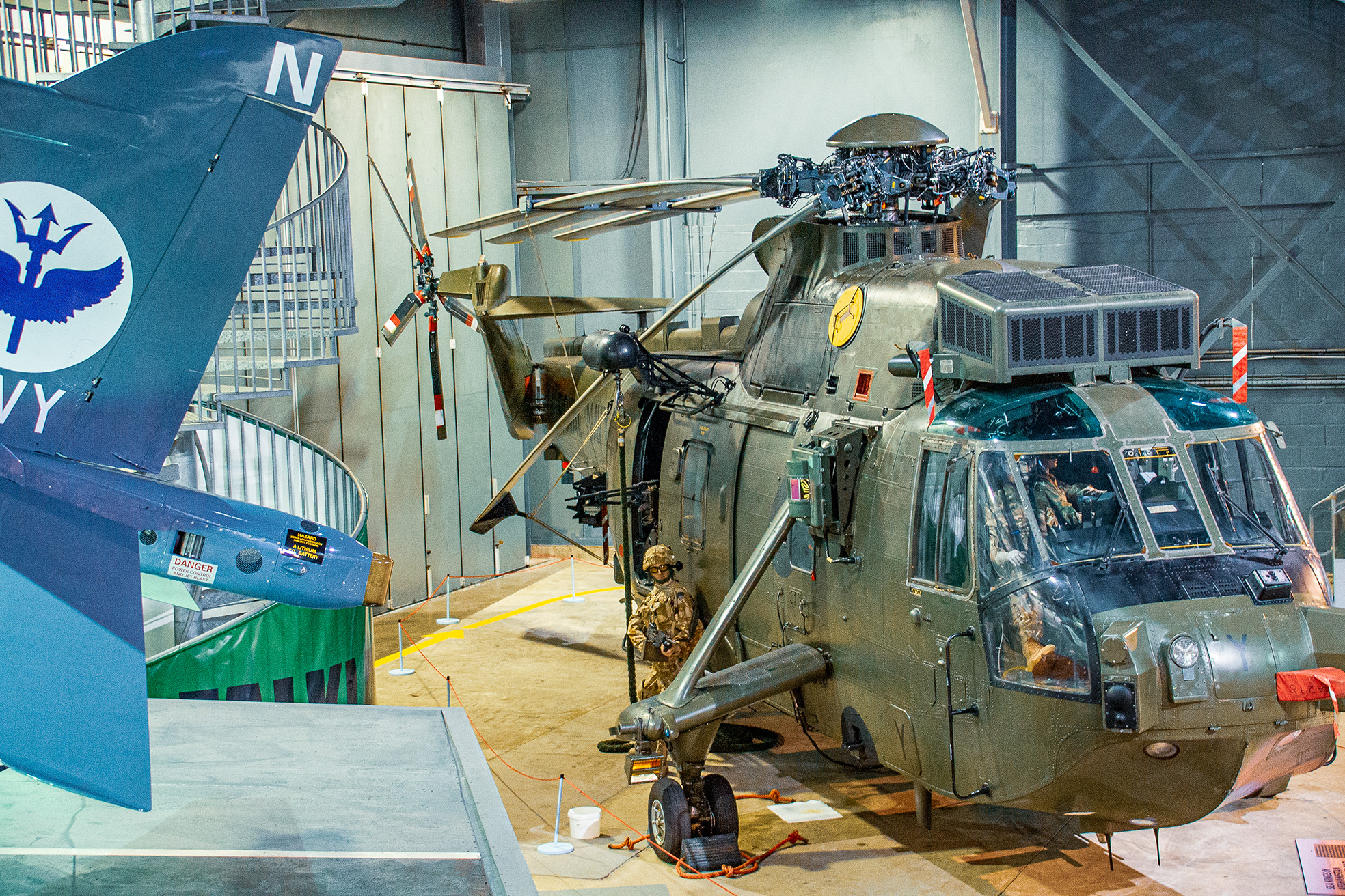 The image shows a military helicopter on display in a museum or exhibition setting. The helicopter is painted in a camouflage pattern and has various equipment attached to it. There is a mannequin dressed in military gear positioned near the helicopter, likely representing a crew member. The tail of the helicopter features a large blue emblem with the word 'NAVY' and an eagle or bird graphic. The setting appears to be indoors, with the helicopter secured on a platform and surrounded by barriers to keep visitors at a safe distance.