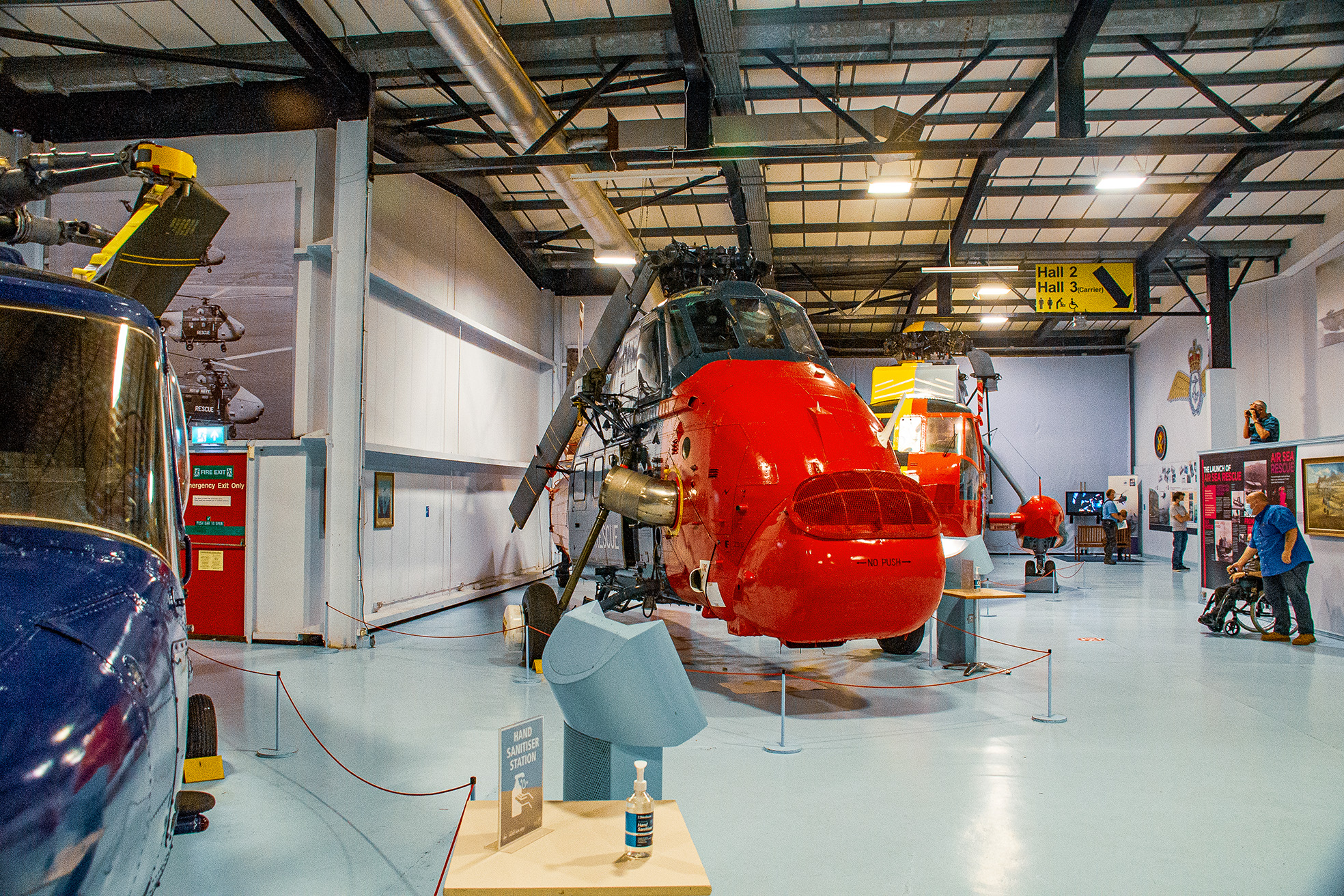 The image depicts an indoor aviation museum exhibit featuring various aircraft, including a prominent red helicopter in the center. The museum has a high ceiling with exposed beams and industrial lighting. Visitors are seen observing the exhibits, and there are informational displays and signs, including a hand sanitizer station in the foreground. The environment suggests a well-organized and educational space dedicated to showcasing historical and technological aspects of aviation.
