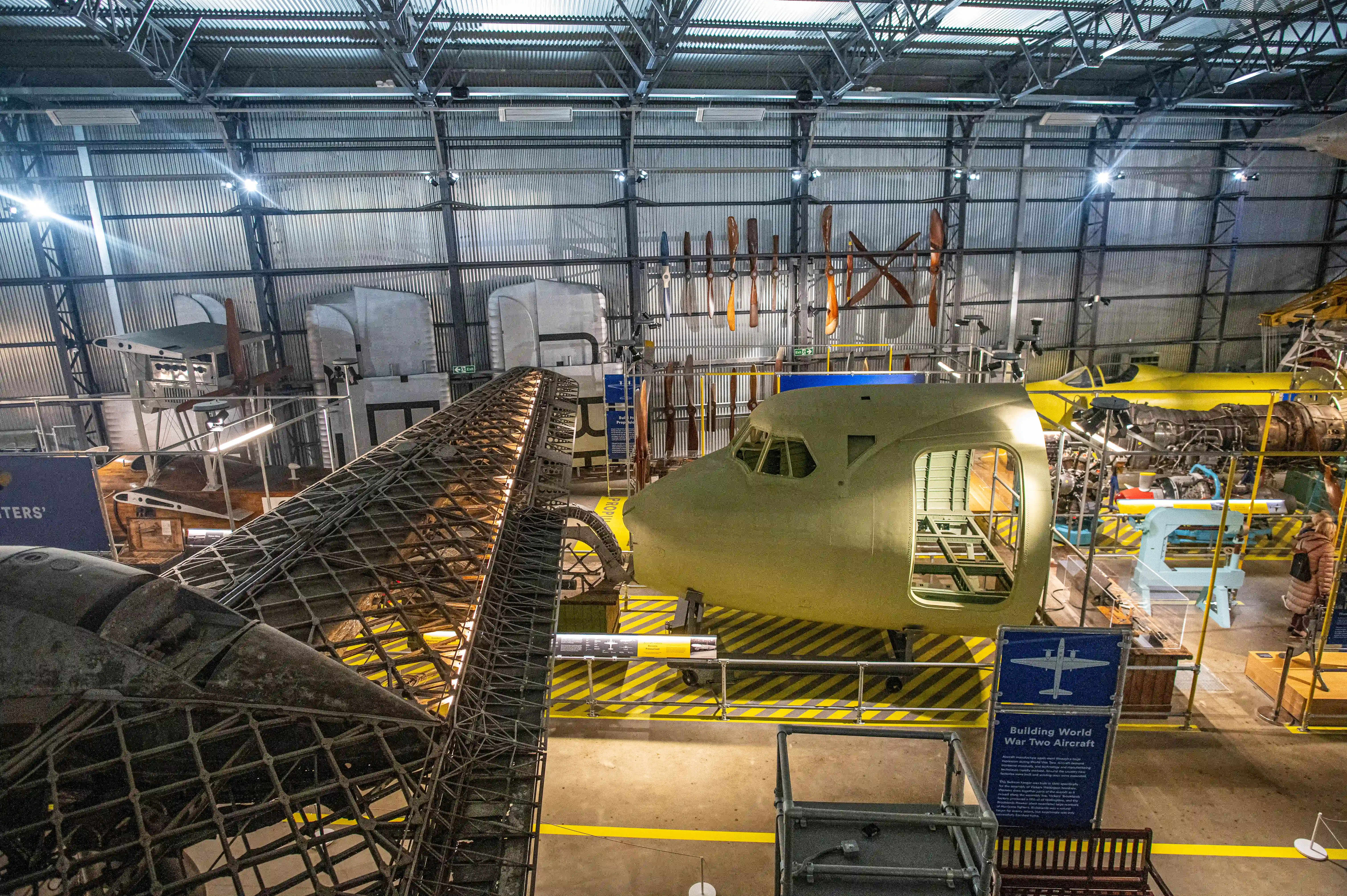 A wide shot of the interior of an aircraft museum hangar, with a prominent light-yellow aircraft fuselage in the foreground, unfinished wing structures, and various vintage aircraft engines and propellers displayed around the space.