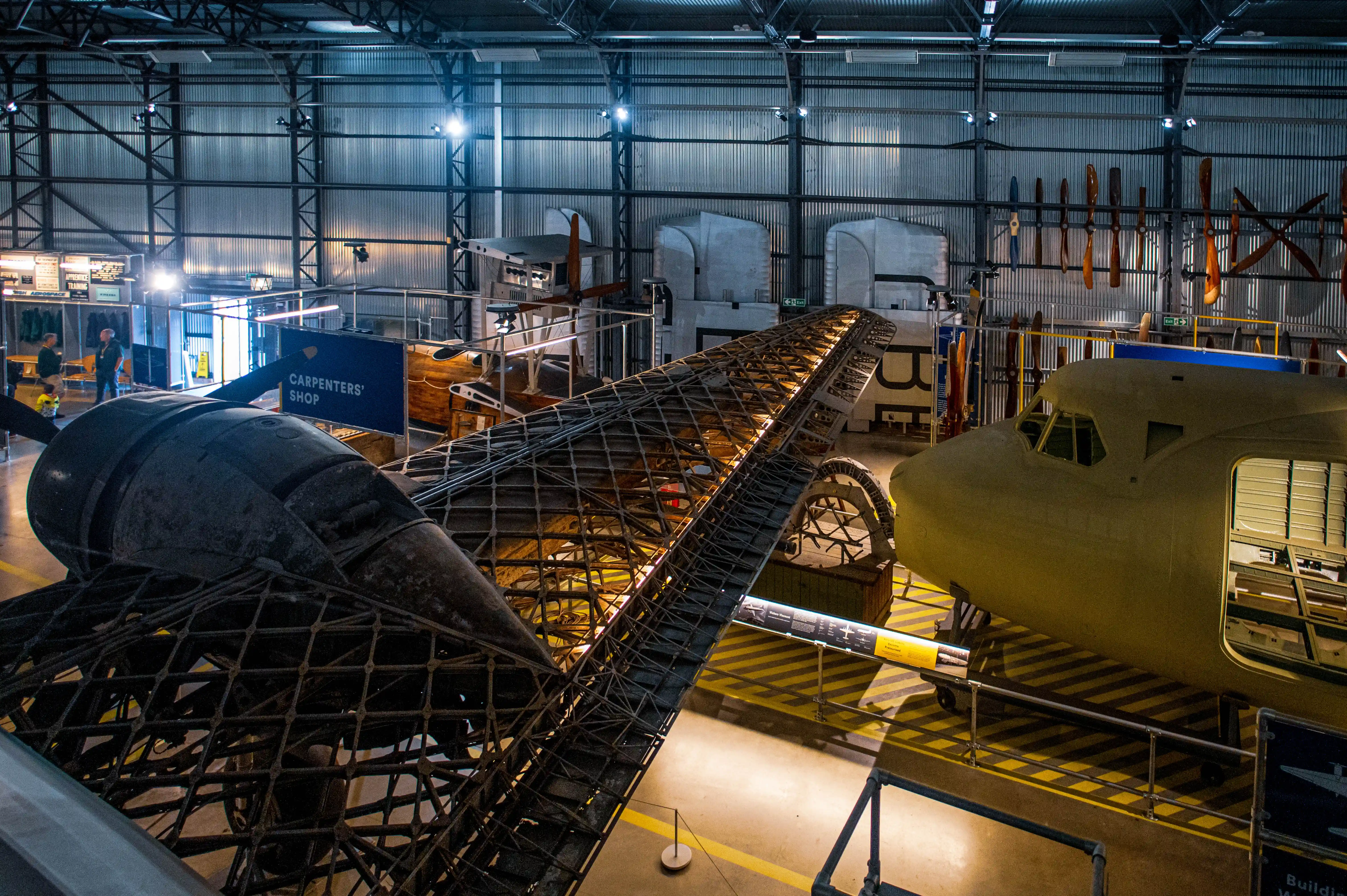 An indoor exhibition space with the exposed metal frame of an aircraft wing angled across the shot. A yellow aircraft fuselage sits to the right, and several vintage propellers are mounted on the corrugated metal wall behind.