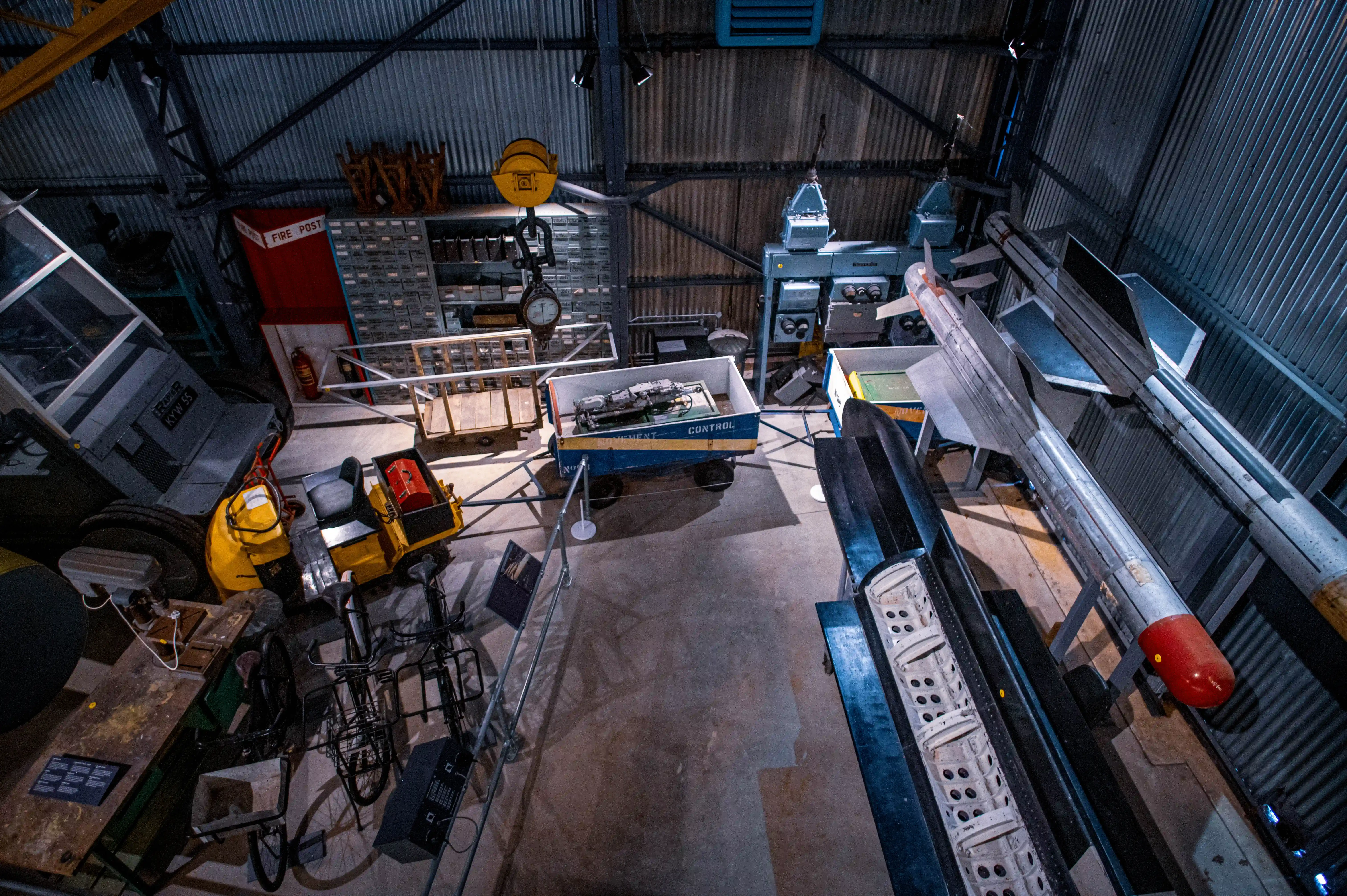 An overhead shot of a museum exhibit featuring a silver missile with a red tip, alongside other military equipment and support vehicles, all displayed within a large industrial building.