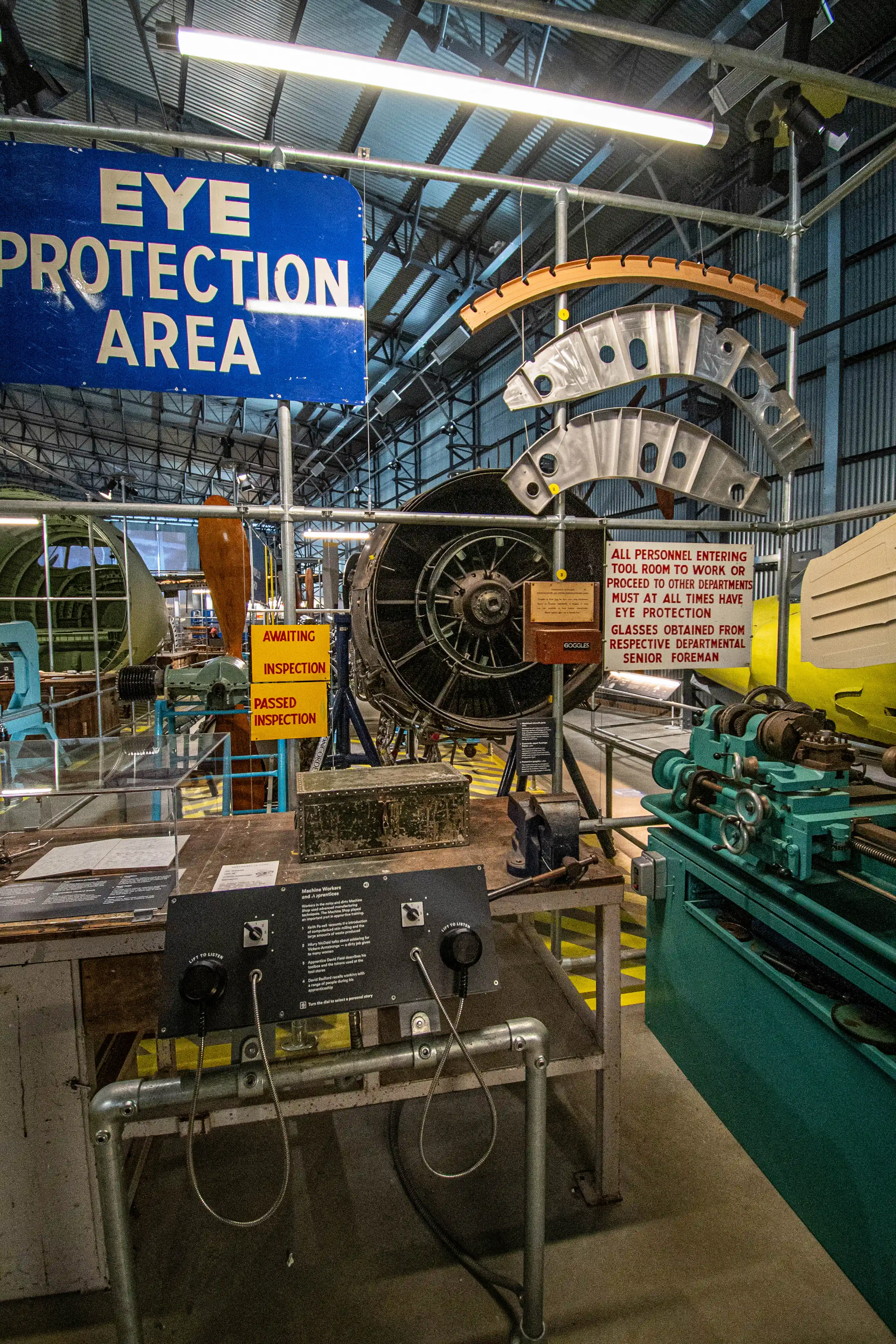A detailed view of historical aircraft engines, A well-lit indoor exhibit depicting an aircraft workshop, with a large jet engine, a teal-coloured lathe, workbenches, and safety signs including EYE PROTECTION AREA and ALL PERSONNEL ENTERING TOOL ROOM TO WORK OR PROCEED TO OTHER DEPARTMENTS MUST AT ALL TIMES HAVE EYE PROTECTION.