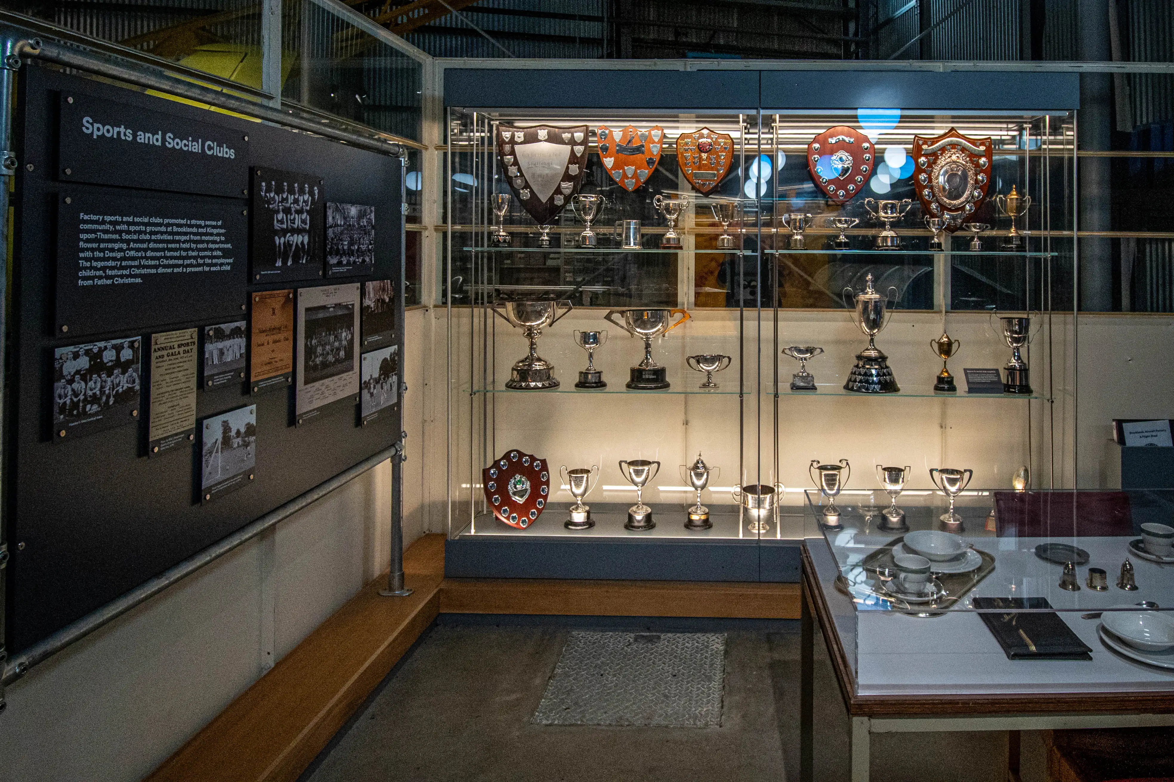 A display case filled with numerous trophies and shields, alongside framed photographs and documents related to factory sports and social clubs, is shown in a museum setting. A descriptive panel details the role of these clubs in promoting community and social activities.
