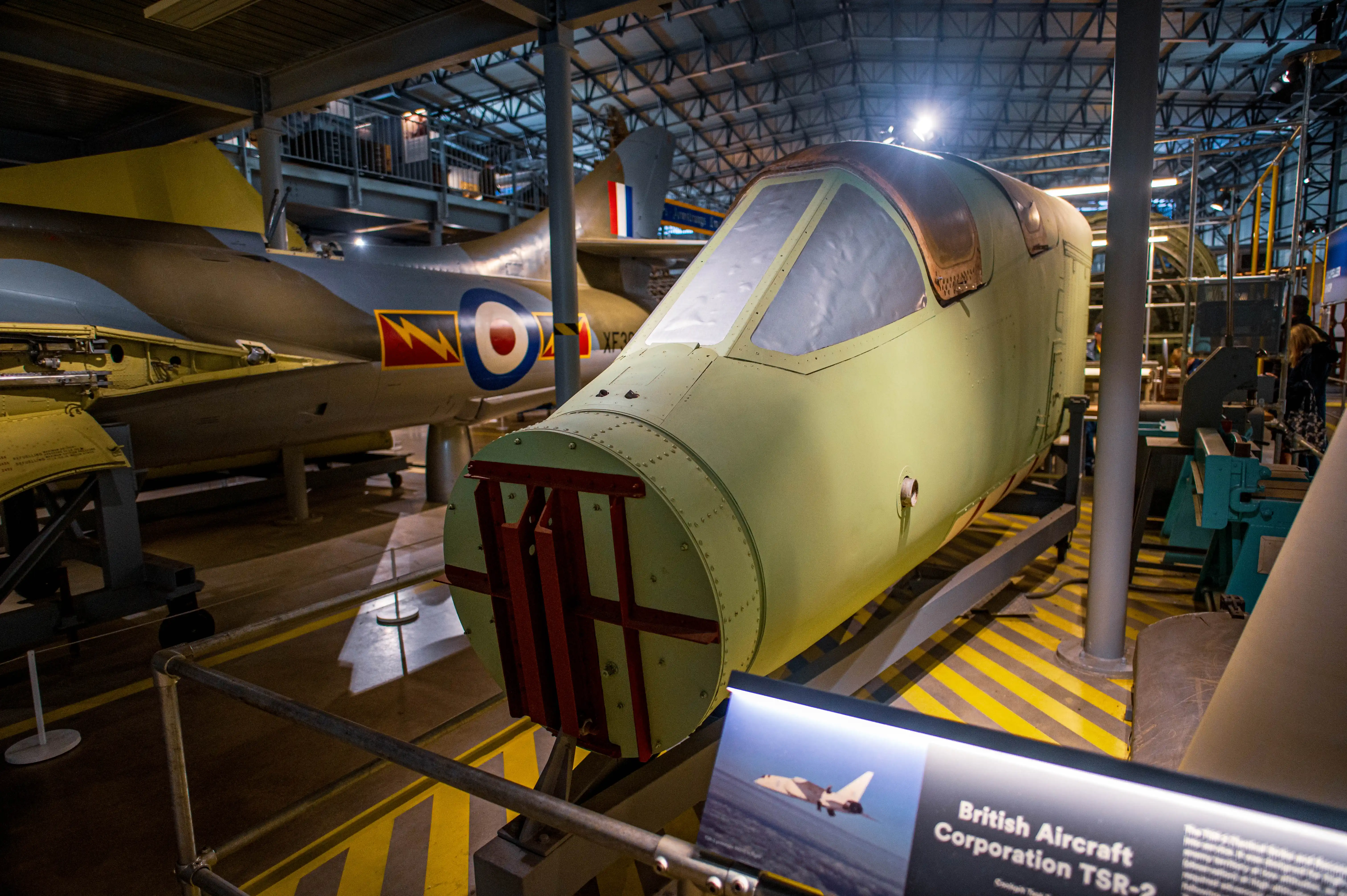 The front section of a pale green military jet aircraft cockpit with a large canopy, displayed indoors. Another aircraft with Royal Air Force roundels is visible in the background.