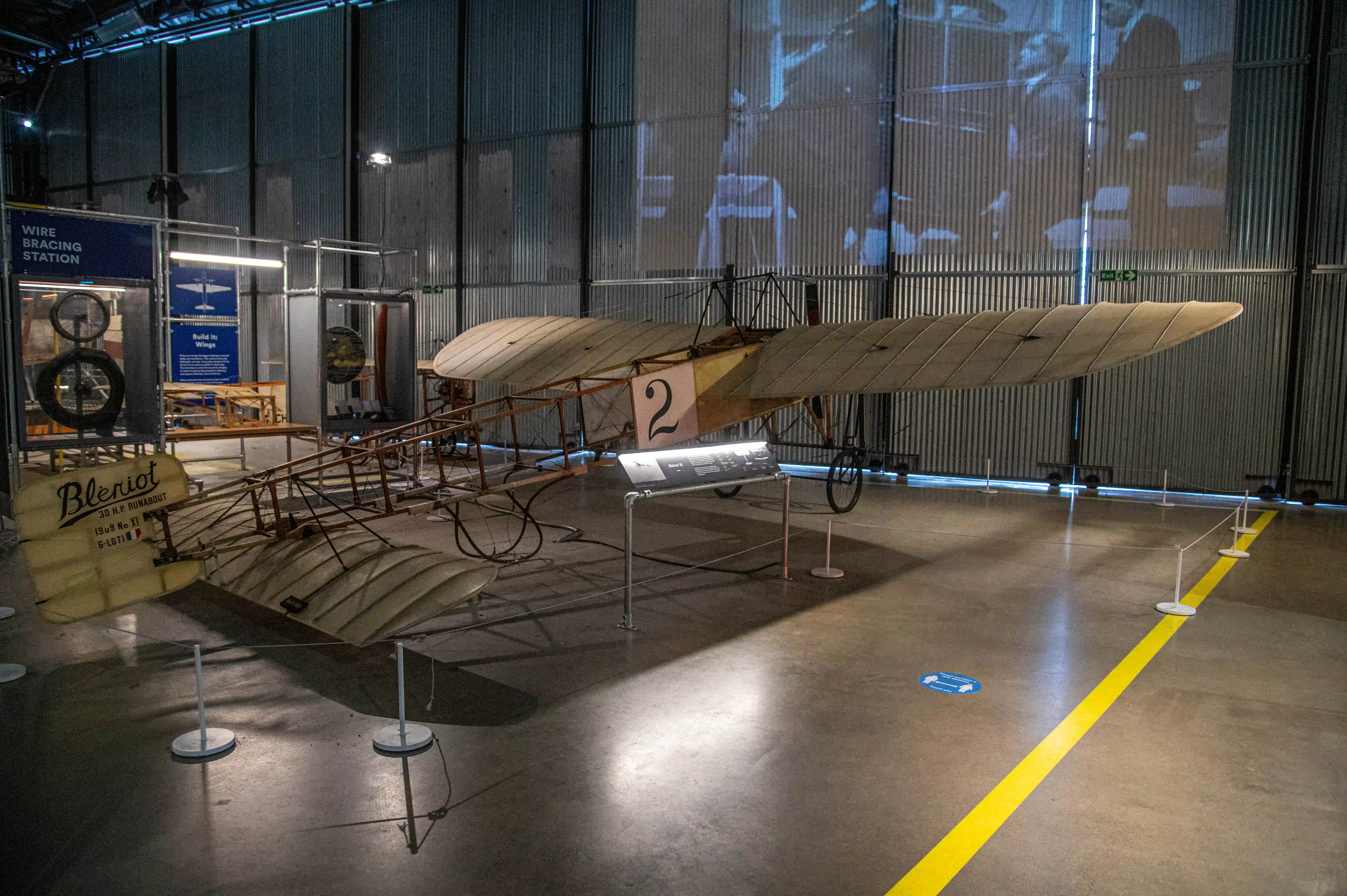 Alt-text: A vintage Blériot XI aeroplane with fabric-covered wings and an open frame sits on display in a museum, with interpretive panels nearby and historical footage projected on the wall behind.