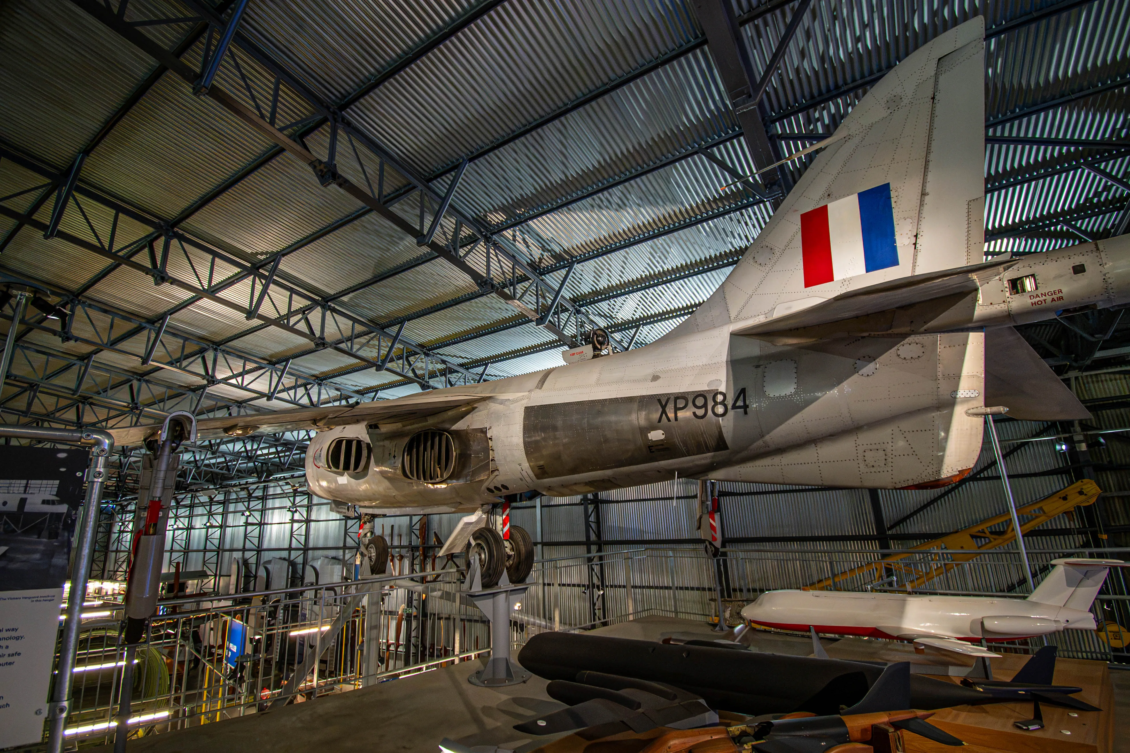 A detailed side view of an Hawker Siddeley P1127 jet aircraft, with registration XP984, displayed in a museum. The aircraft's tail fin displays the red, white, and blue Royal Air Force roundel. It is suspended in a hangar with other exhibits visible below.