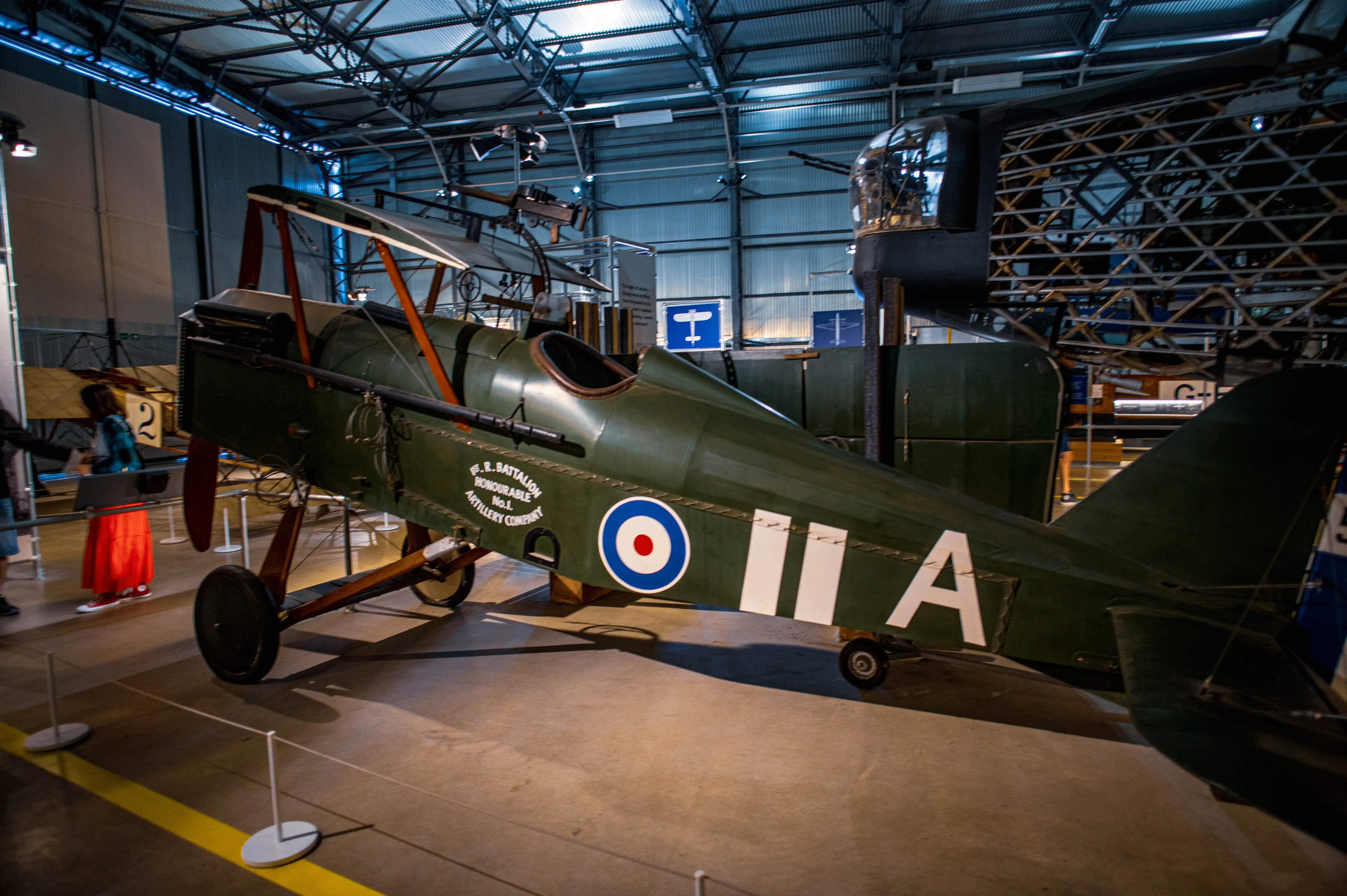 A dark green World War I era biplane is on display in a museum hangar. The aircraft has a single cockpit, wooden propeller, and is marked with a circular RAF roundel. Other exhibits are visible in the background.