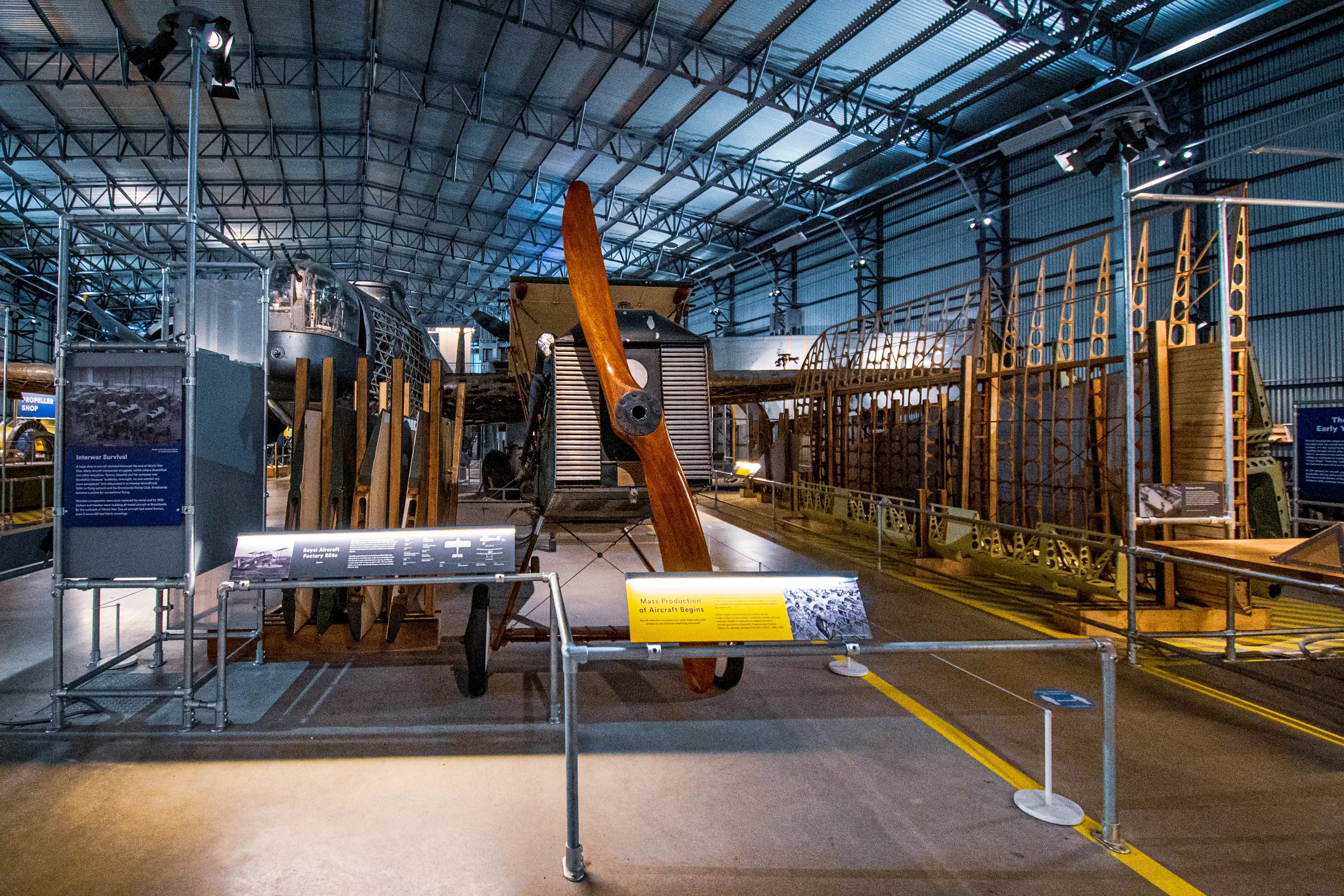 Interior of a museum hangar displaying the skeletal framework of aircraft and a large wooden propeller, with interpretive signs detailing early aviation history.