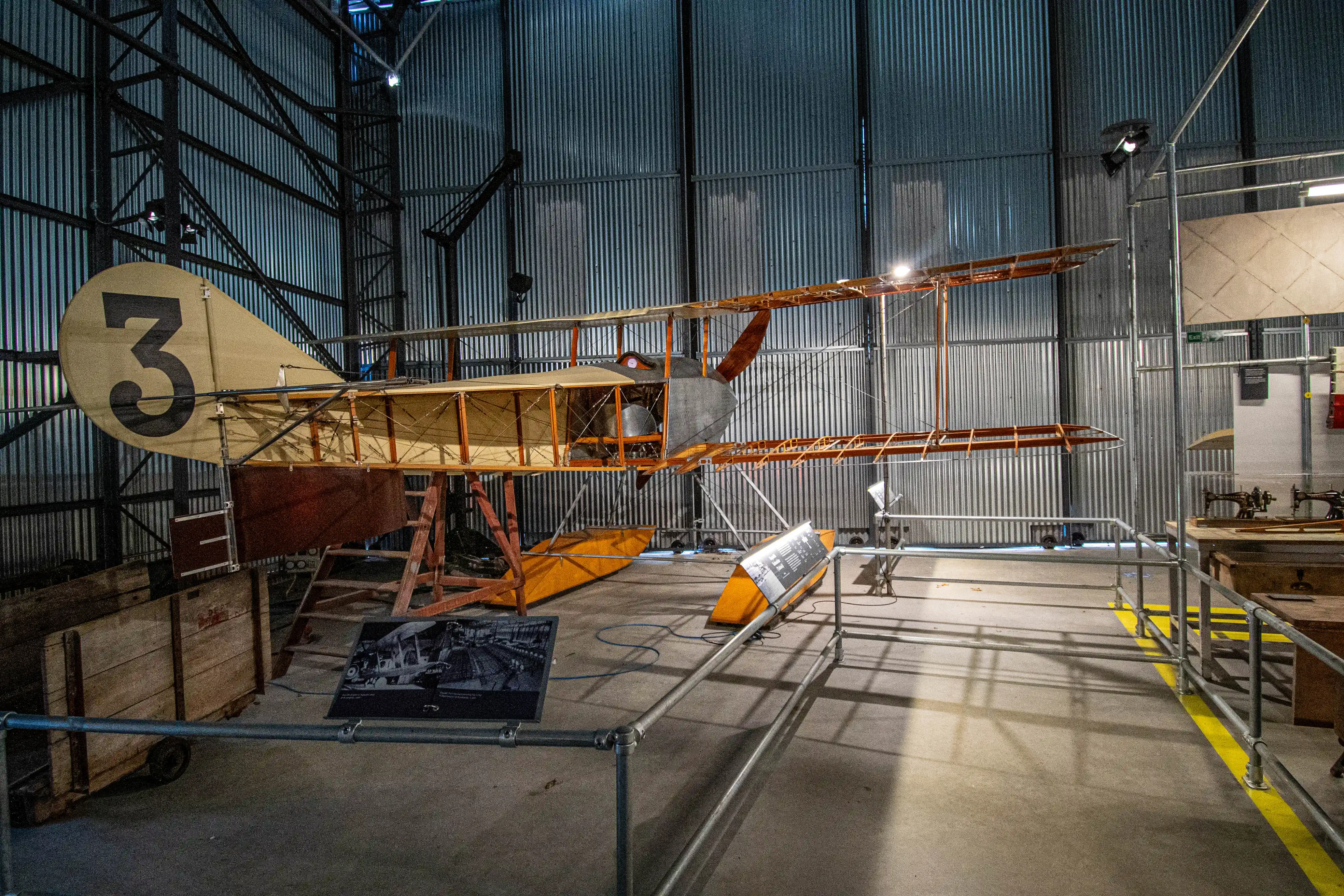 A vintage biplane, identified by a large black number '3' on its tail, is showcased within the corrugated metal walls of an aviation museum. The aircraft, supported by wooden scaffolding, features a fabric-covered fuselage and wings, with visible internal wiring and bracing. Informative display panels are positioned near the exhibit, along with a wooden crate and a section of yellow safety flooring.