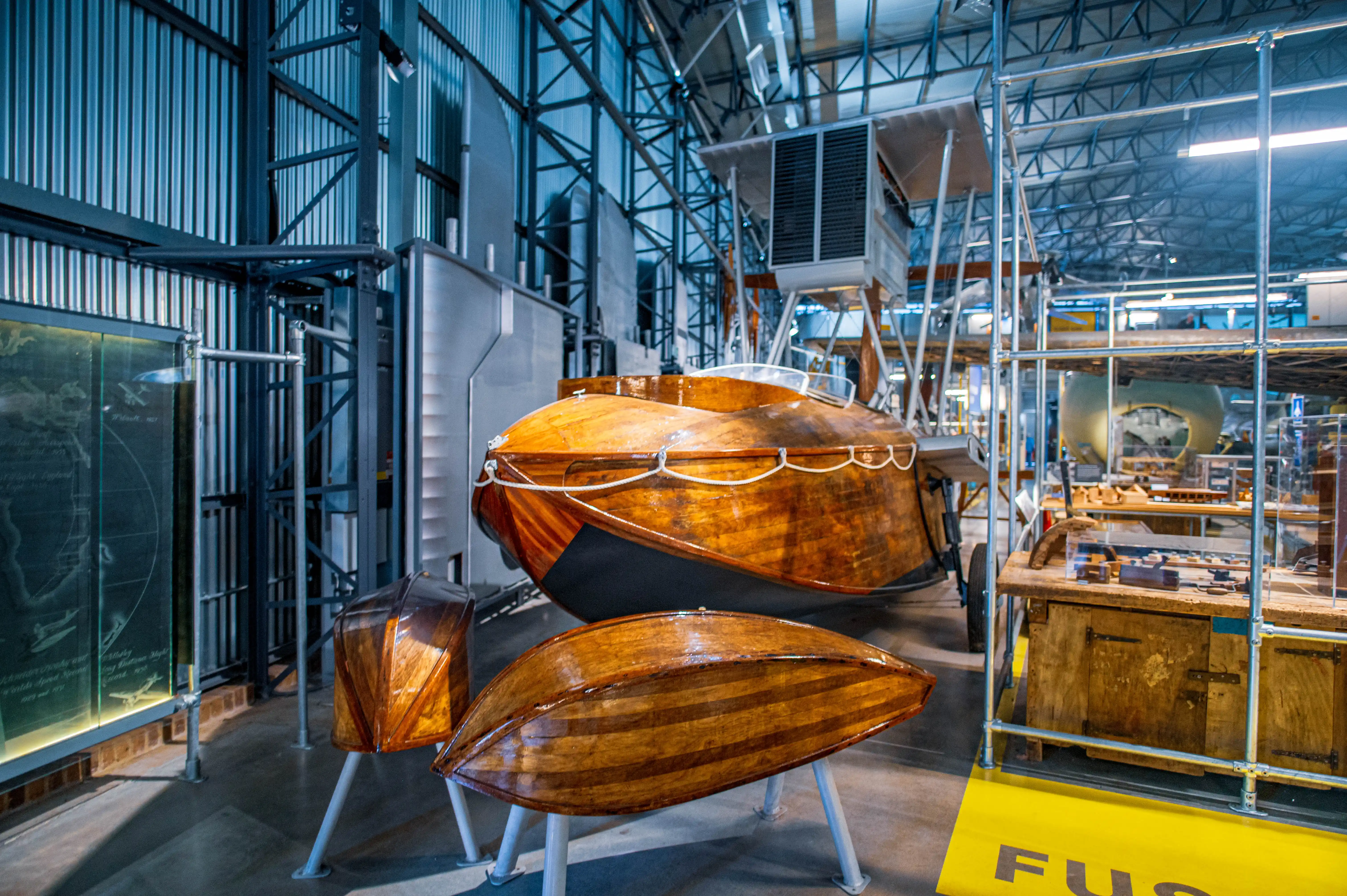 Polished wooden aircraft fuselage suspended in a museum with smaller varnished wooden components displayed beneath it.