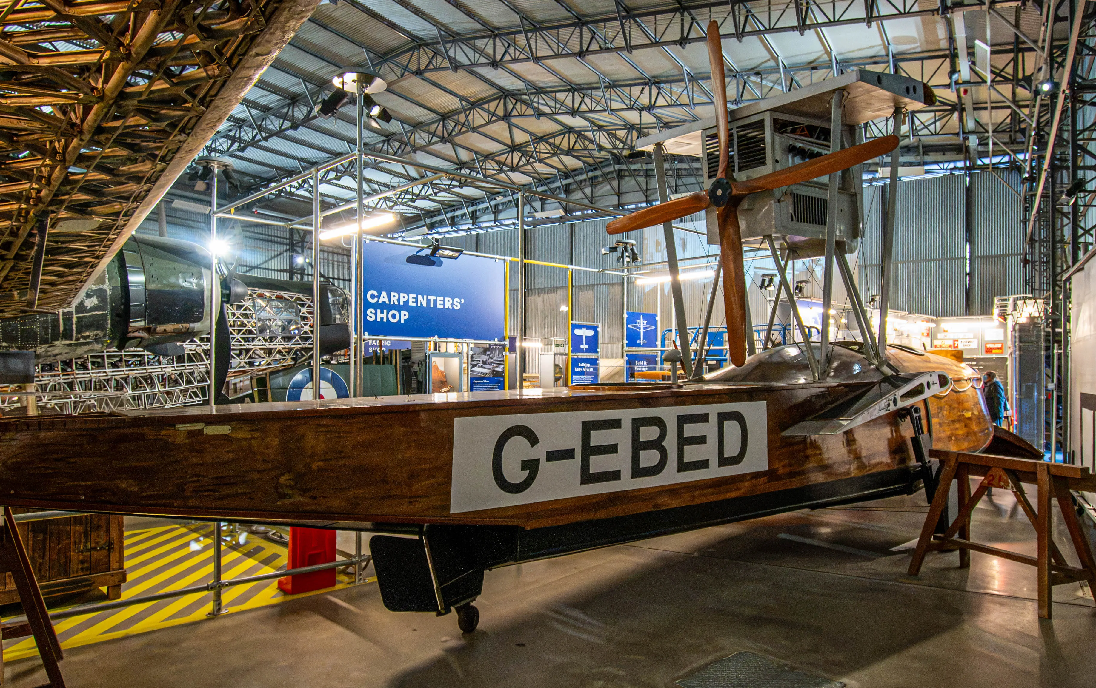  A vintage biplane, identified by the registration G-EBED, is exhibited indoors within a museum setting. The aircraft, primarily constructed from wood and fabric, features a prominent propeller. In the background, a sign reads CARPENTERS' SHOP, suggesting an area dedicated to the restoration or construction of aircraft. Other exhibits and structural elements of the hangar are also visible.