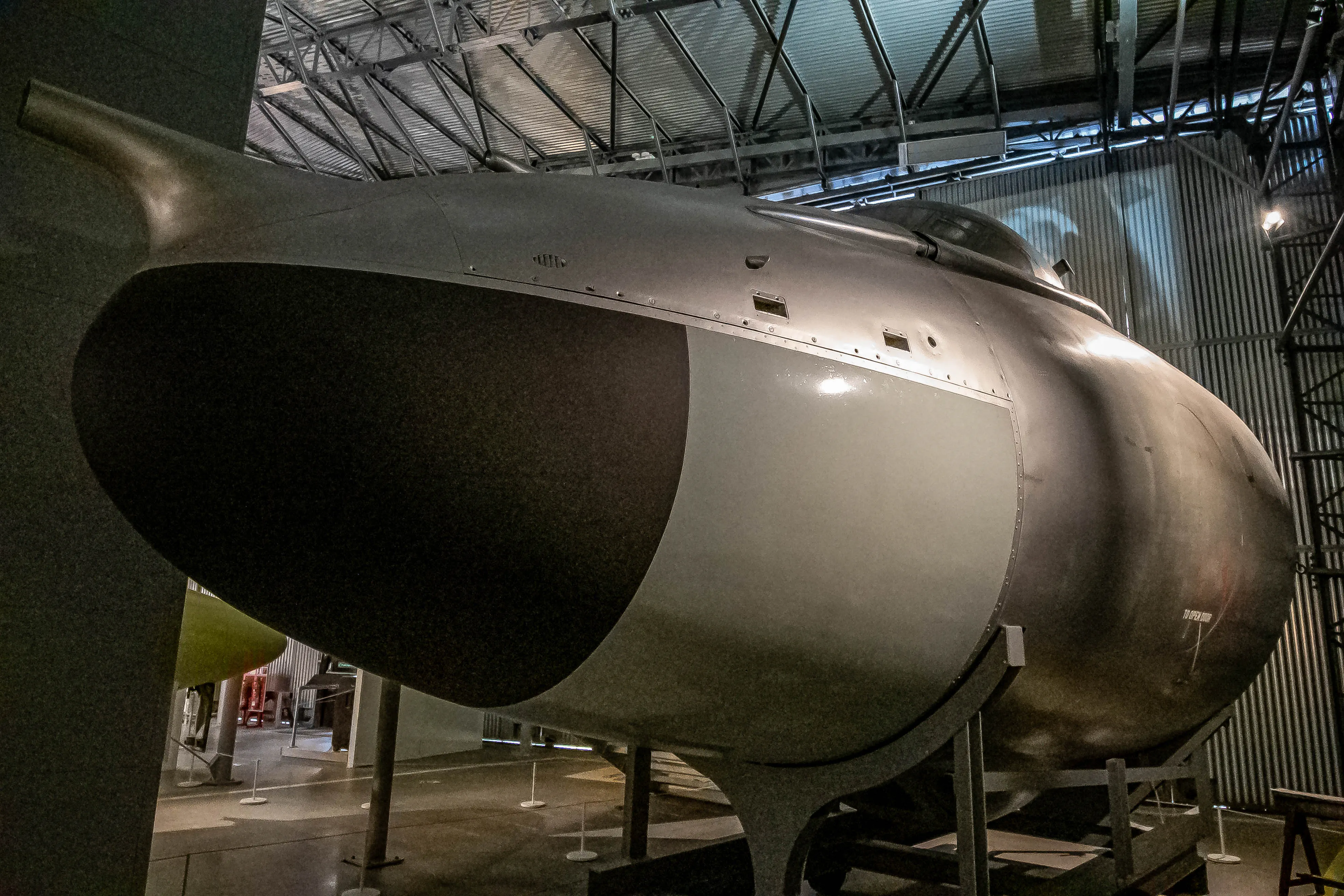 An aerial view of the front section of a silver aeroplane with a dark grey cockpit window area, displayed indoors. A person stands to the right of the aircraft, looking at it.