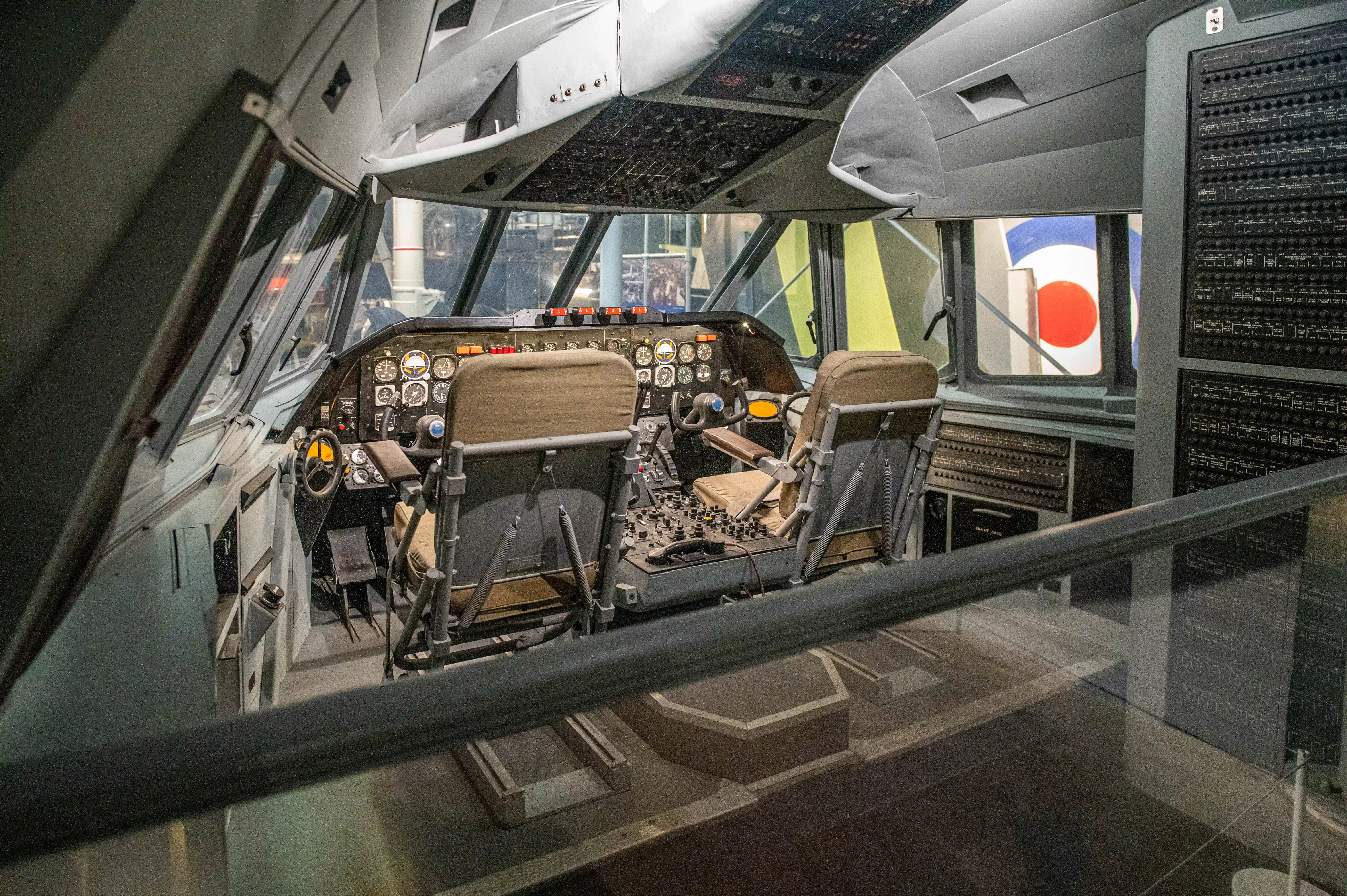 Interior view of an old aeroplane cockpit, featuring two pilot seats, steering yokes, an array of analogue gauges and switches, and a window displaying a red and blue circular aircraft marking.