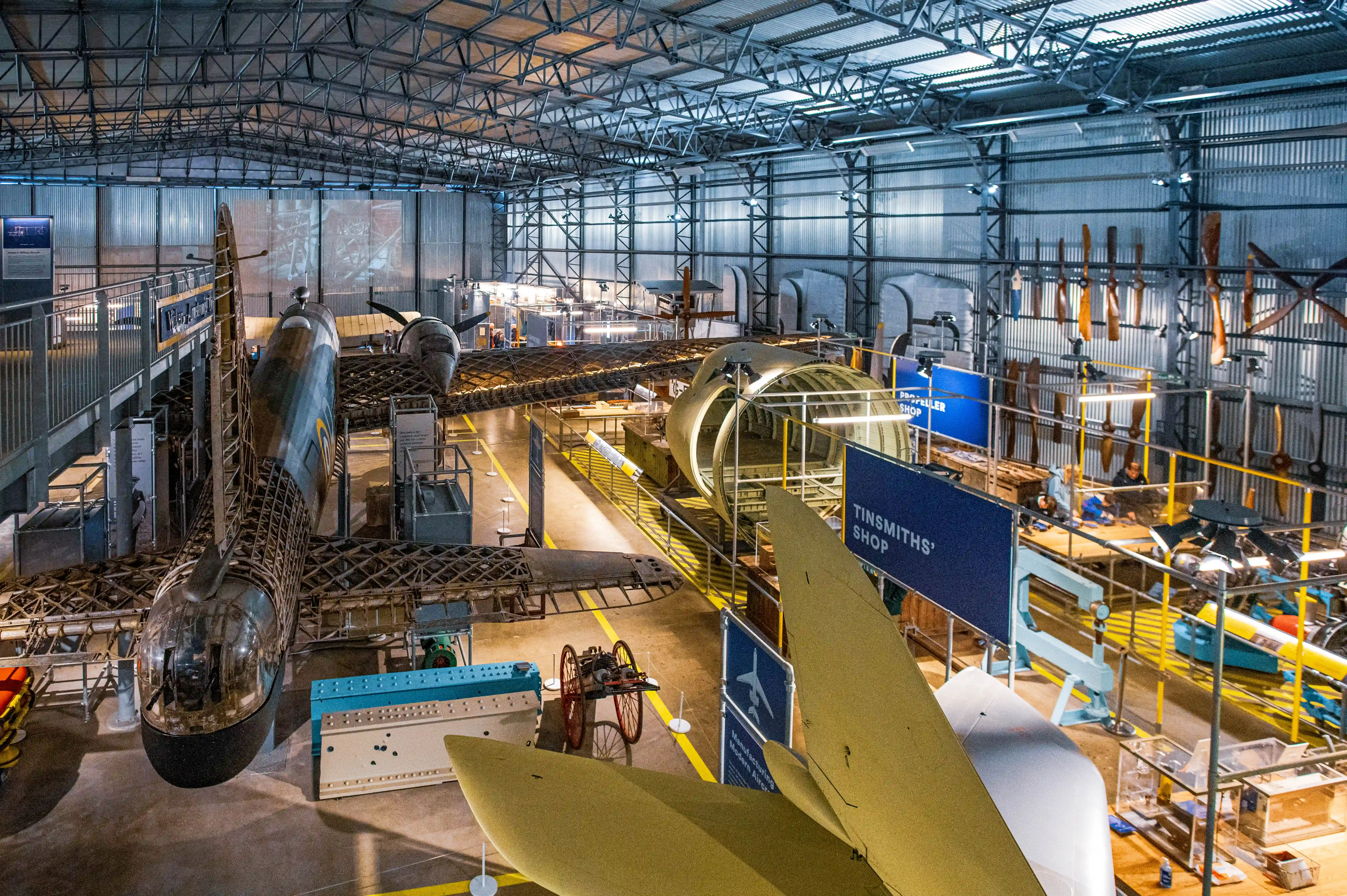  Interior of an aviation museum showing a partially assembled aircraft suspended overhead, with displays of aircraft parts, tools, and historical aviation exhibits.