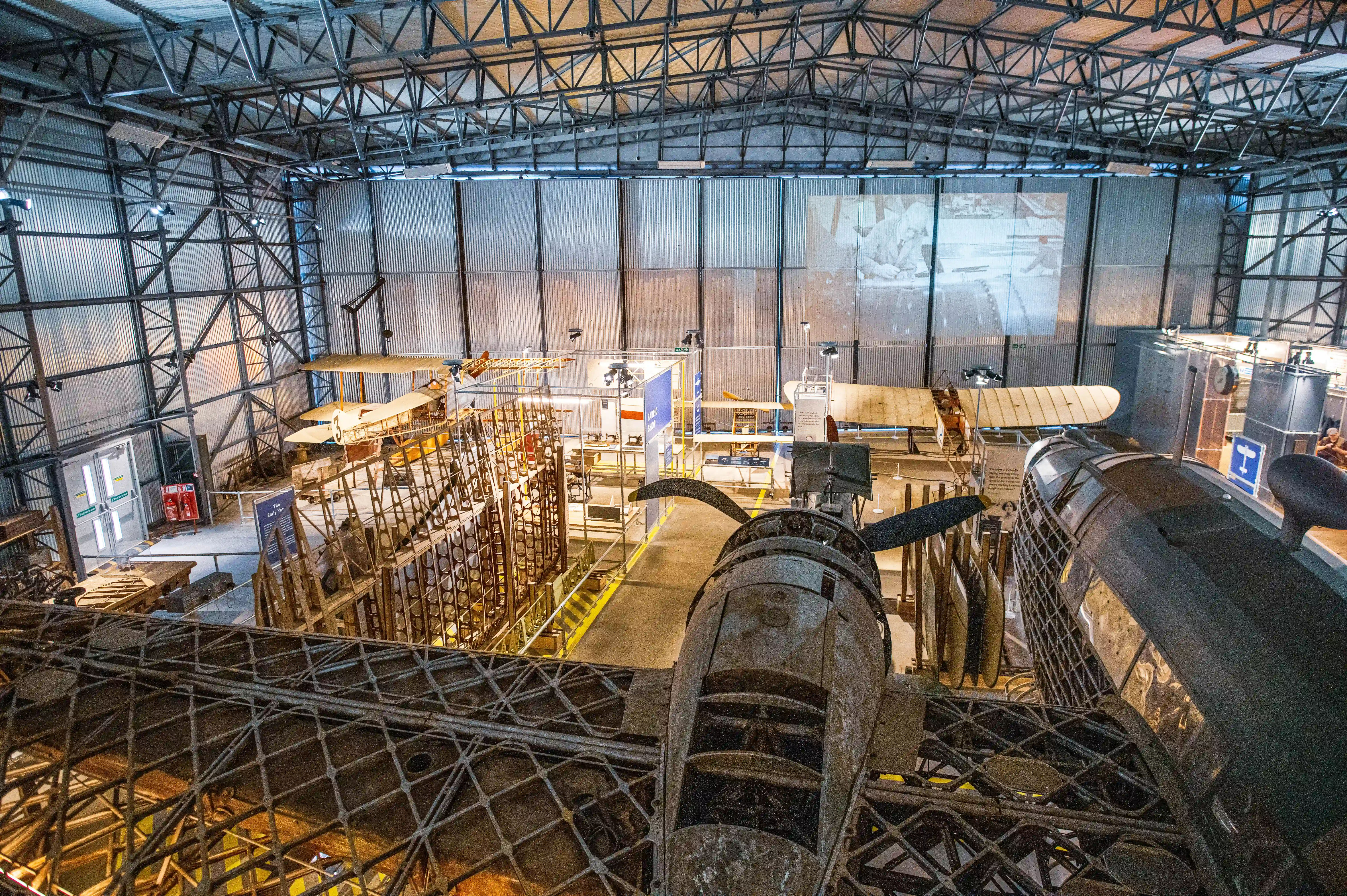 Interior view of an aviation museum with multiple vintage aircraft, including a biplane and parts of a bomber in the foreground, displayed within a large industrial hangar.