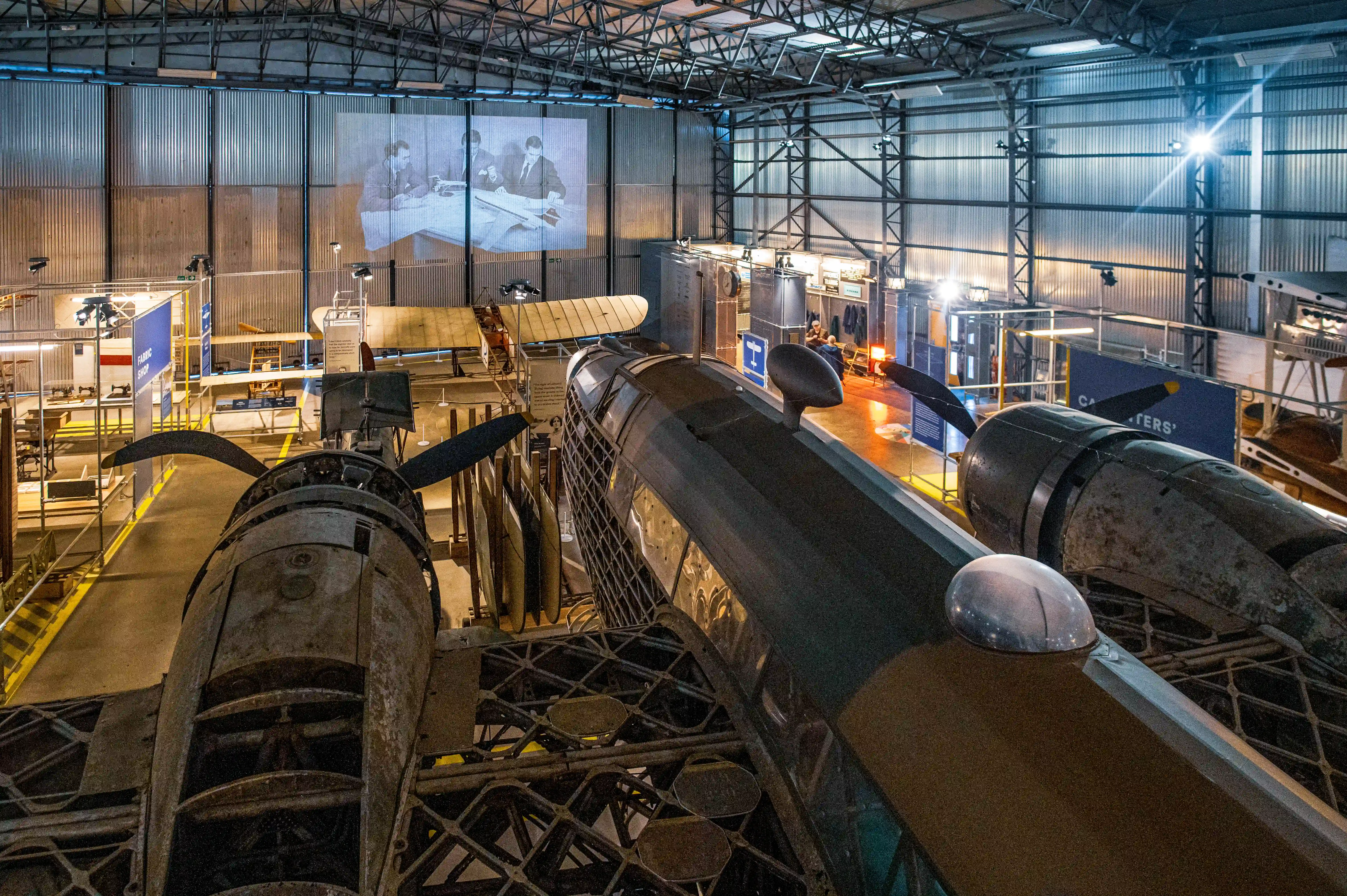 Overhead view of a museum hangar showcasing a vintage bomber aircraft. A projected image of a historical meeting is visible on the back wall, with displays and other aircraft parts arranged around the central exhibit.