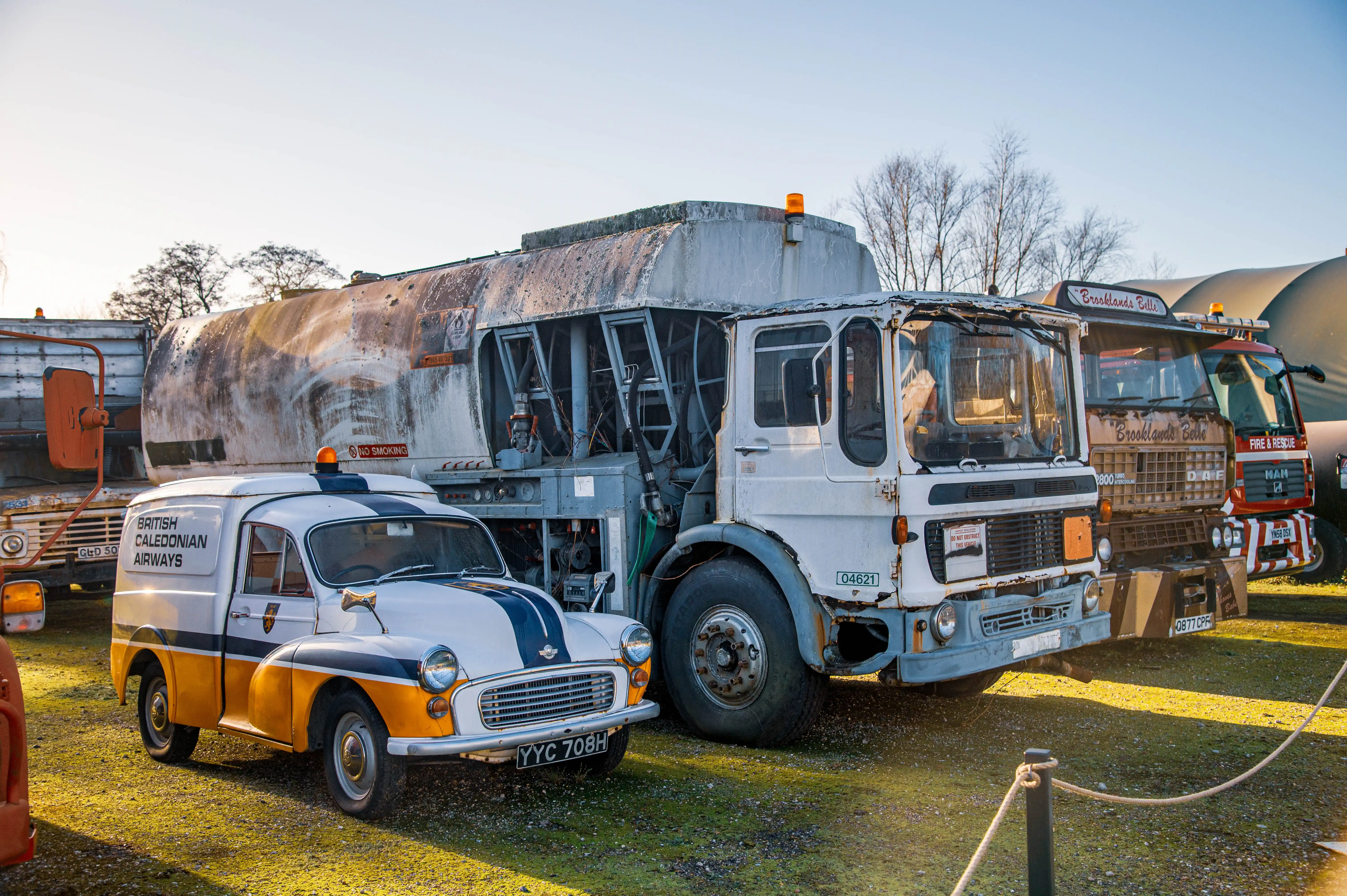 A vintage white and yellow van with BRITISH CALEDONIAN AIRWAYS lettering is parked in front of a large, weathered tanker truck. Behind them, two other trucks, one labelled Brooklands Bells and another with FIRE & RESCUE are visible.