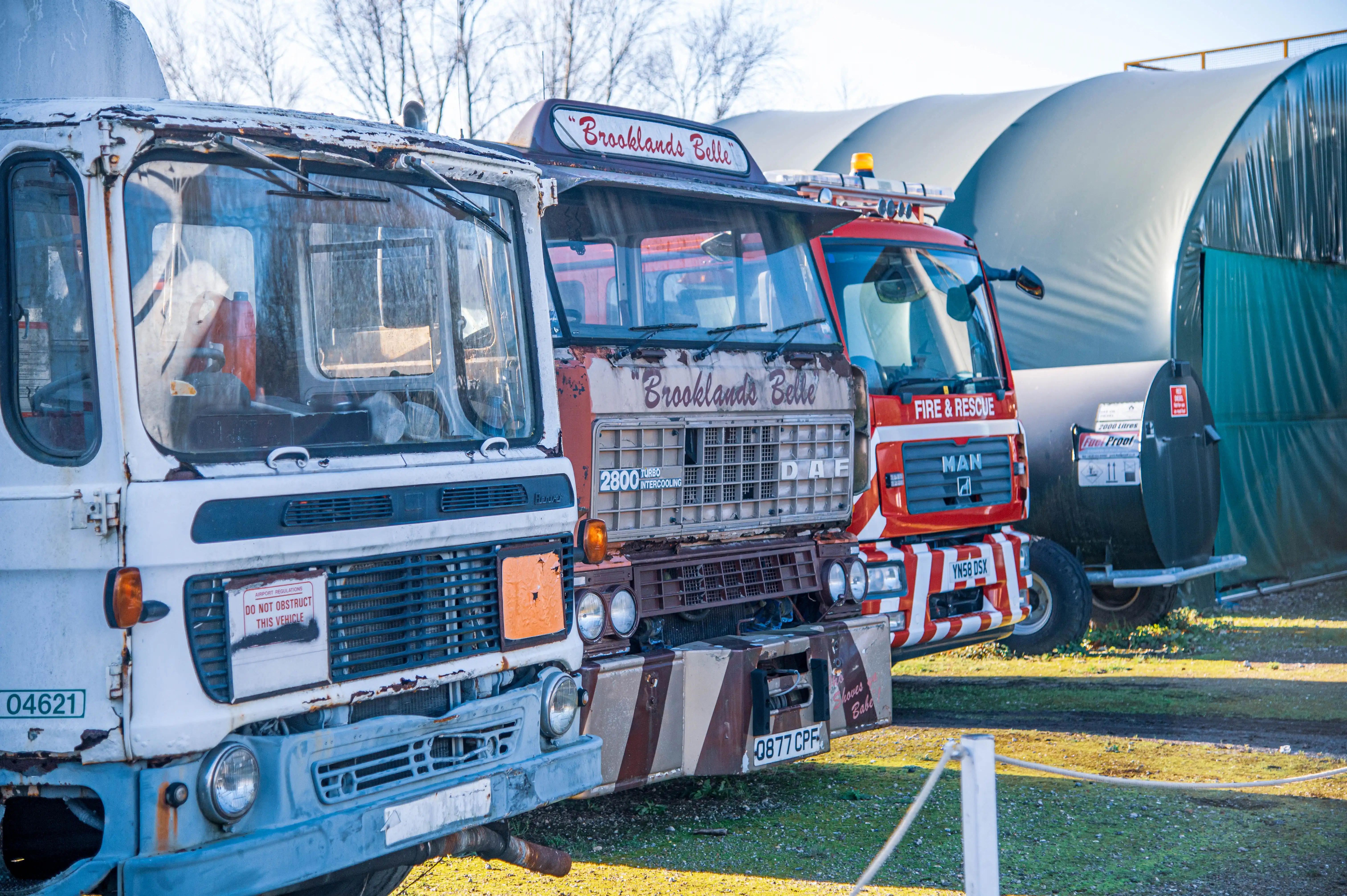 Three trucks lined up, the closest being white, followed by a brown and white truck named Brooklands Belle, and a red and white MAN fire engine, with a large green tent-like structure in the background.