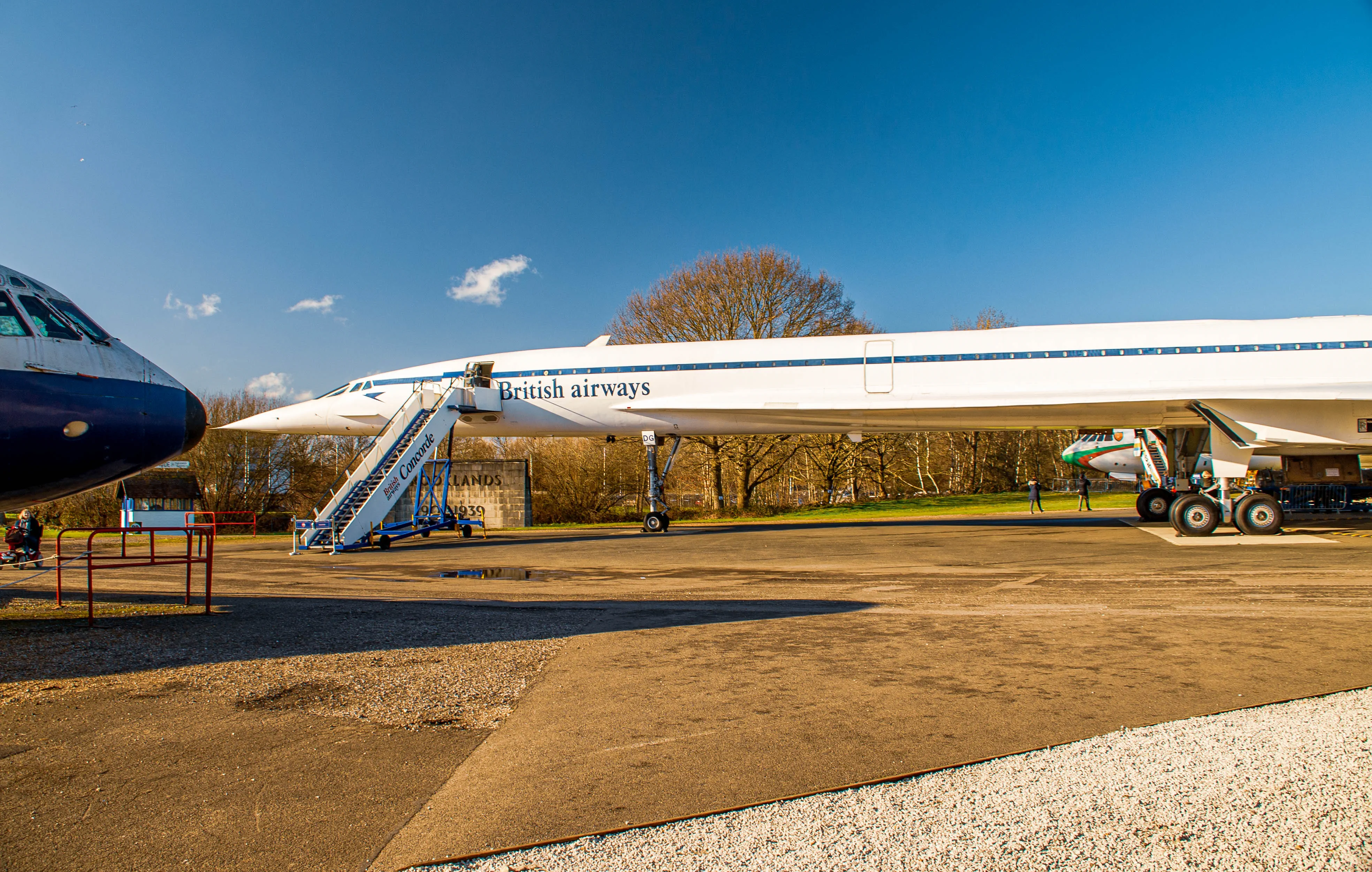 A British Airways Concorde aircraft, painted white with blue accents, sits on an airfield with a set of boarding stairs extended to its door. The nose of another aircraft is visible to the left.