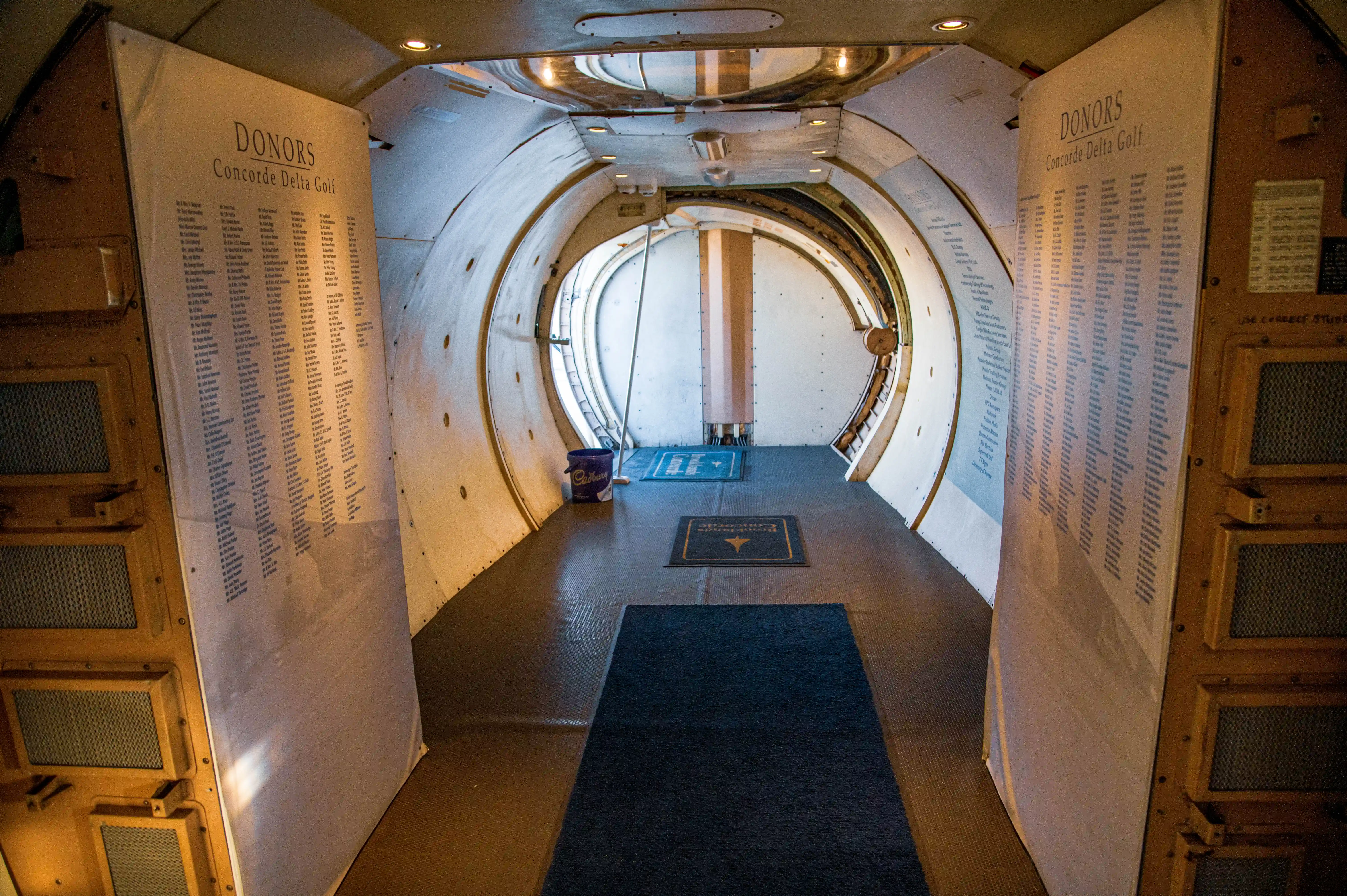 A view down the curved interior corridor of a Concorde aircraft, featuring prominent lists of donors to the Concorde Delta Golf project displayed on large panels. Two welcome mats are placed on the floor.