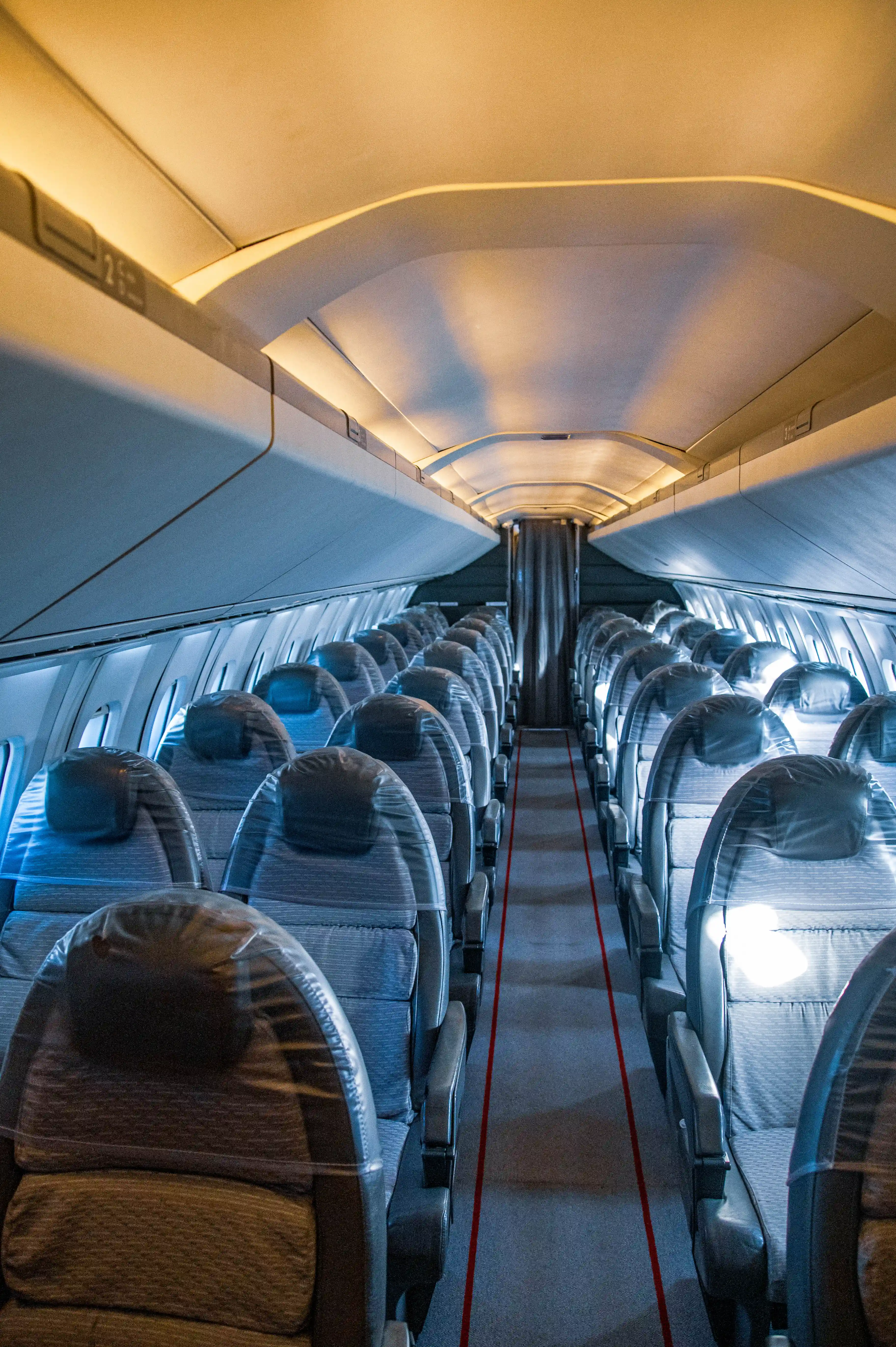 Interior of an aeroplane cabin showing rows of empty seats covered in clear plastic, with an aisle in the centre.