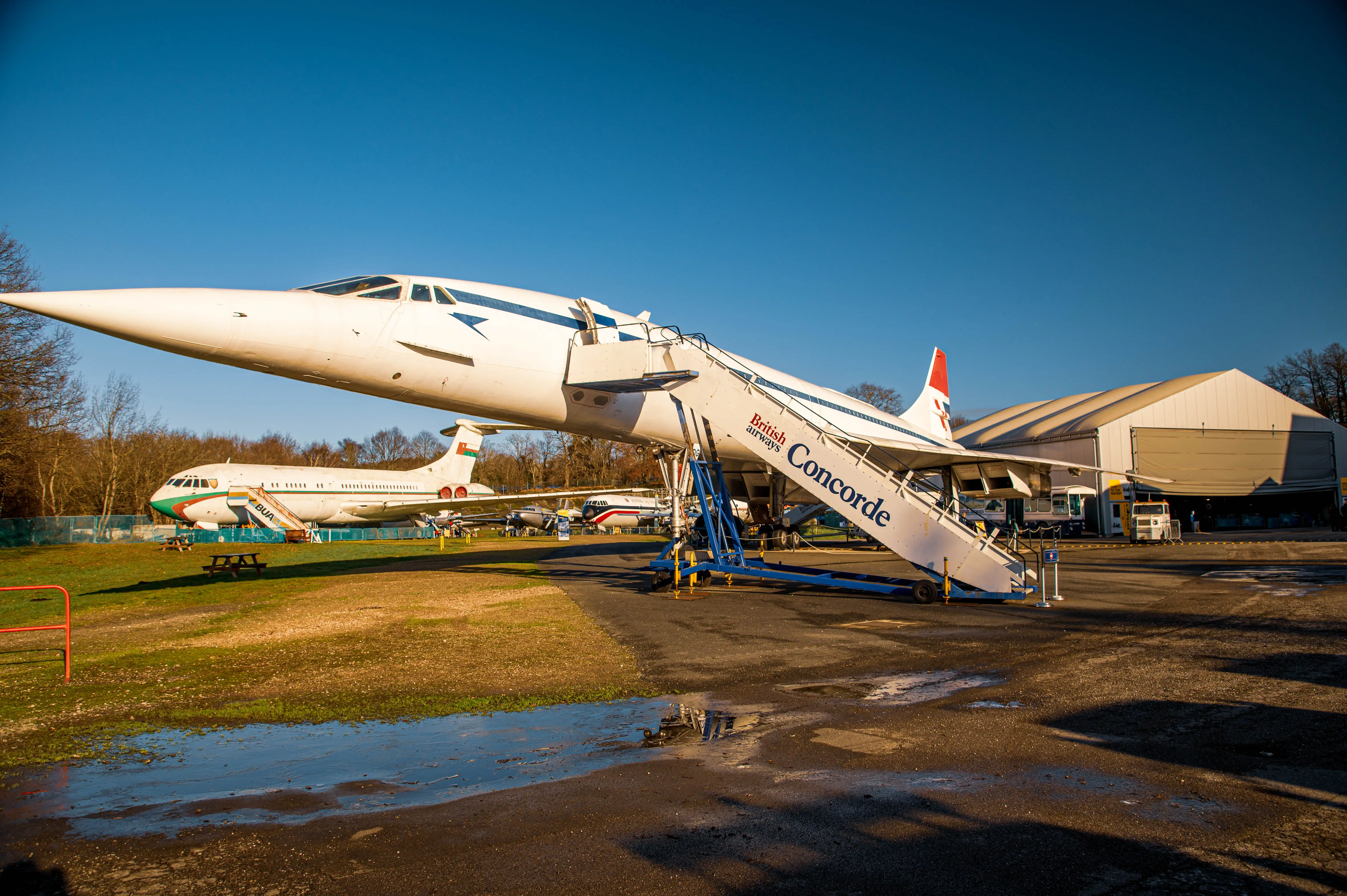 A British Airways Concorde airplane with its iconic delta wing and pointed nose is positioned on the ground, with a mobile stairway attached. In the background, another white aircraft with green and red livery is visible, along with a large white hangar and trees under a bright blue sky.