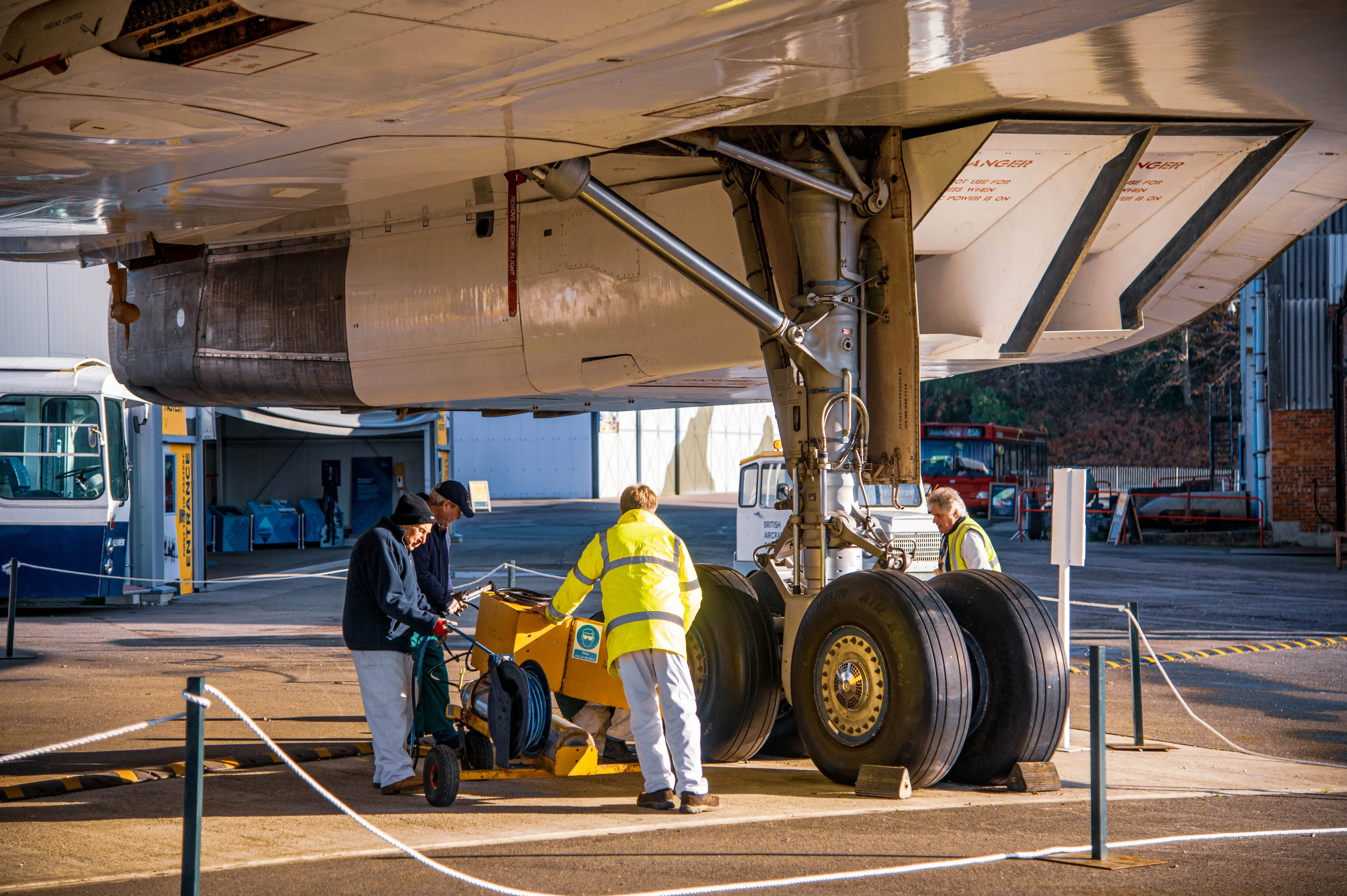 A team of aviation technicians performing maintenance on the landing gear of a large aircraft. The technicians are dressed in high-visibility vests and safety gear, using specialized equipment to inspect or repair the landing gear. The aircraft is stationed in a hangar or maintenance area, with other vehicles and equipment visible in the background.