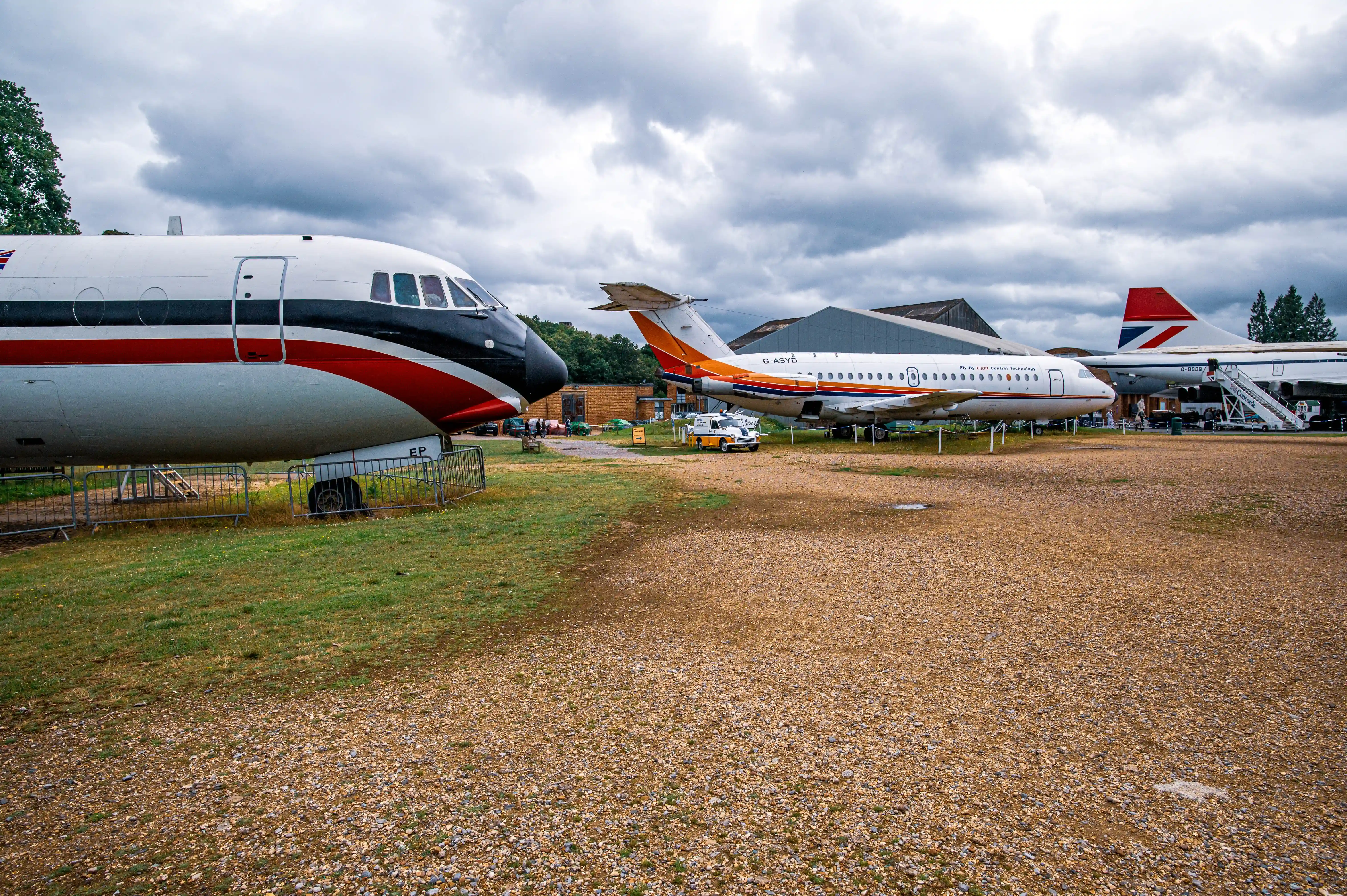 A wide shot of several vintage aeroplanes parked on a gravel and grass area. The closest aeroplane is white with red and black stripes along its side. To its right is a smaller white aeroplane with orange and yellow stripes. In the background is a larger aeroplane with a British Airways tail fin. The sky is cloudy and grey.