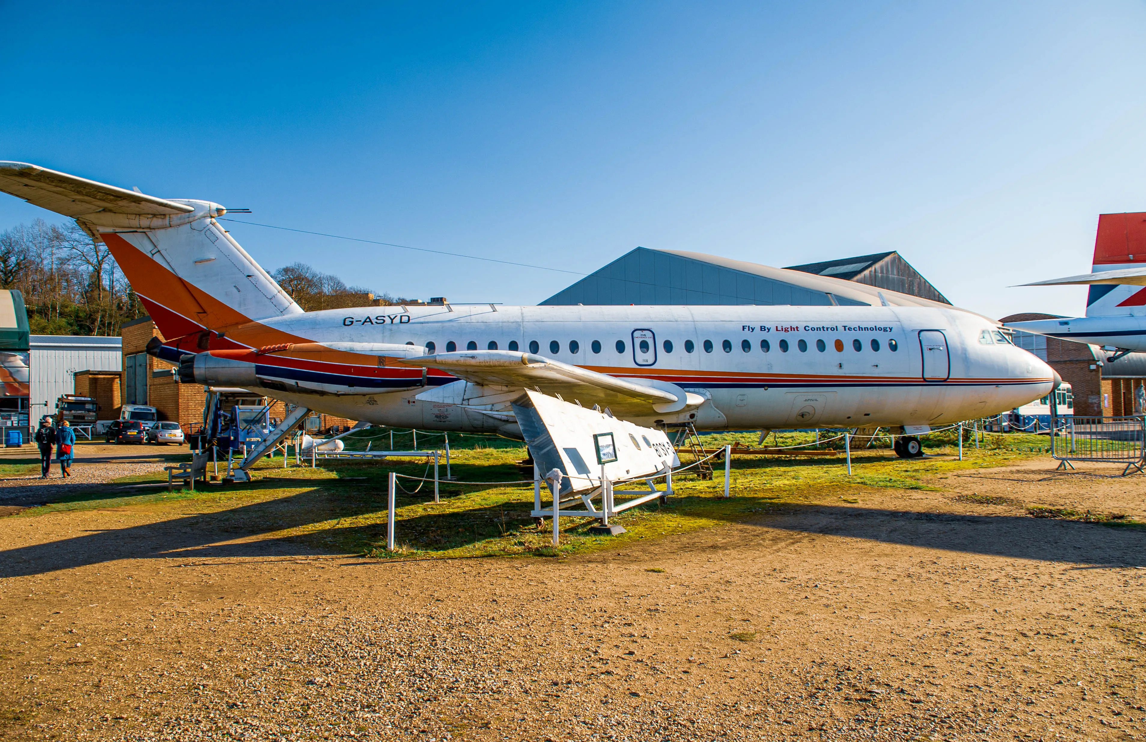 A vintage aircraft with the registration 'G-ASYD' displayed outdoors on a gravel surface. The aircraft has a white fuselage with red and blue stripes, a T-tail design, and jet engines. The phrase 'Fly By Light Control Technology' is visible on the side. In the background, a modern hangar and other aircraft are visible, along with a clear blue sky and trees.