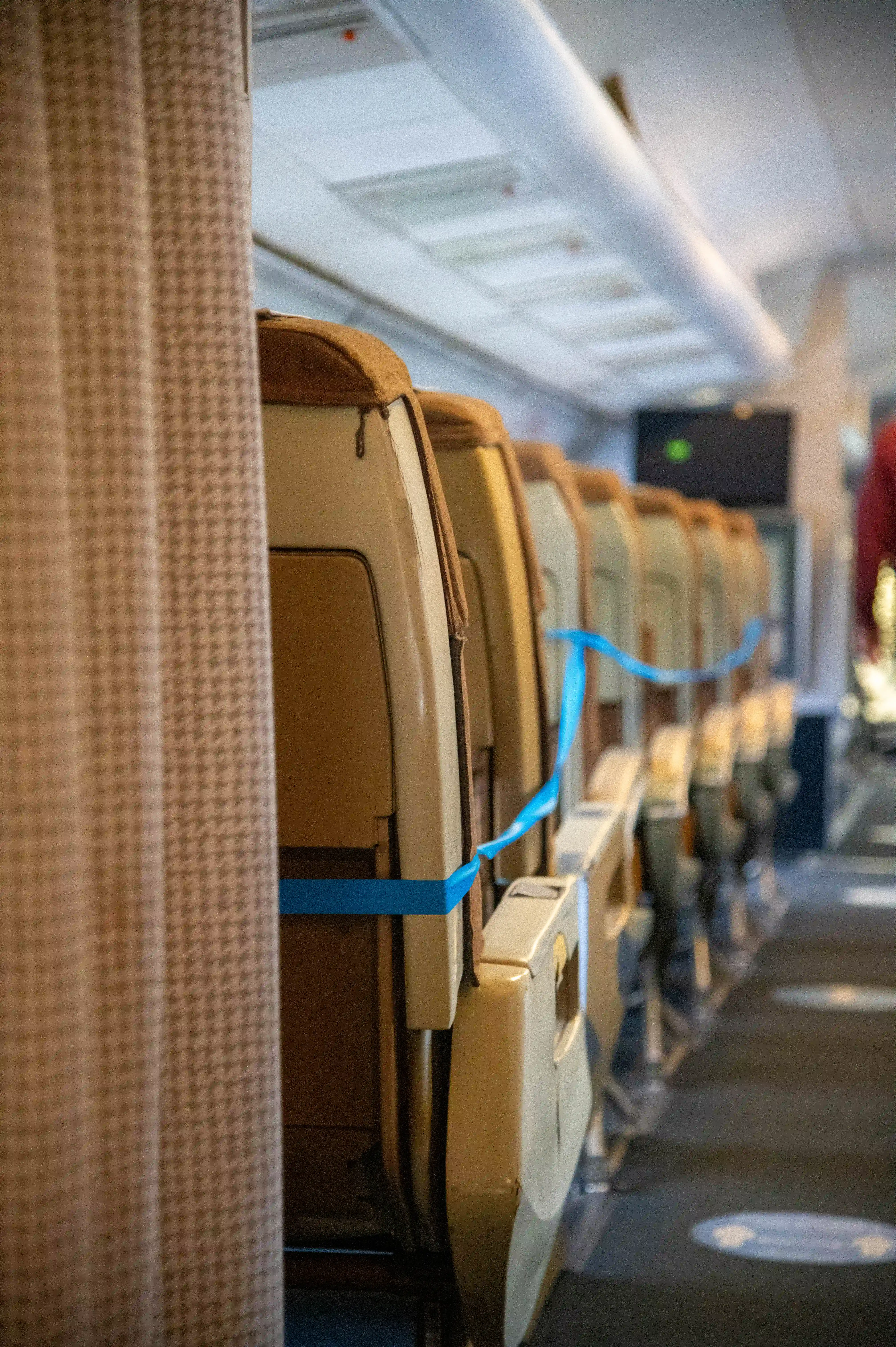  Interior view of an airplane cabin with several empty seats sectioned off by blue tape. A patterned curtain is visible on the left, and social distancing markers are on the floor of the aisle
