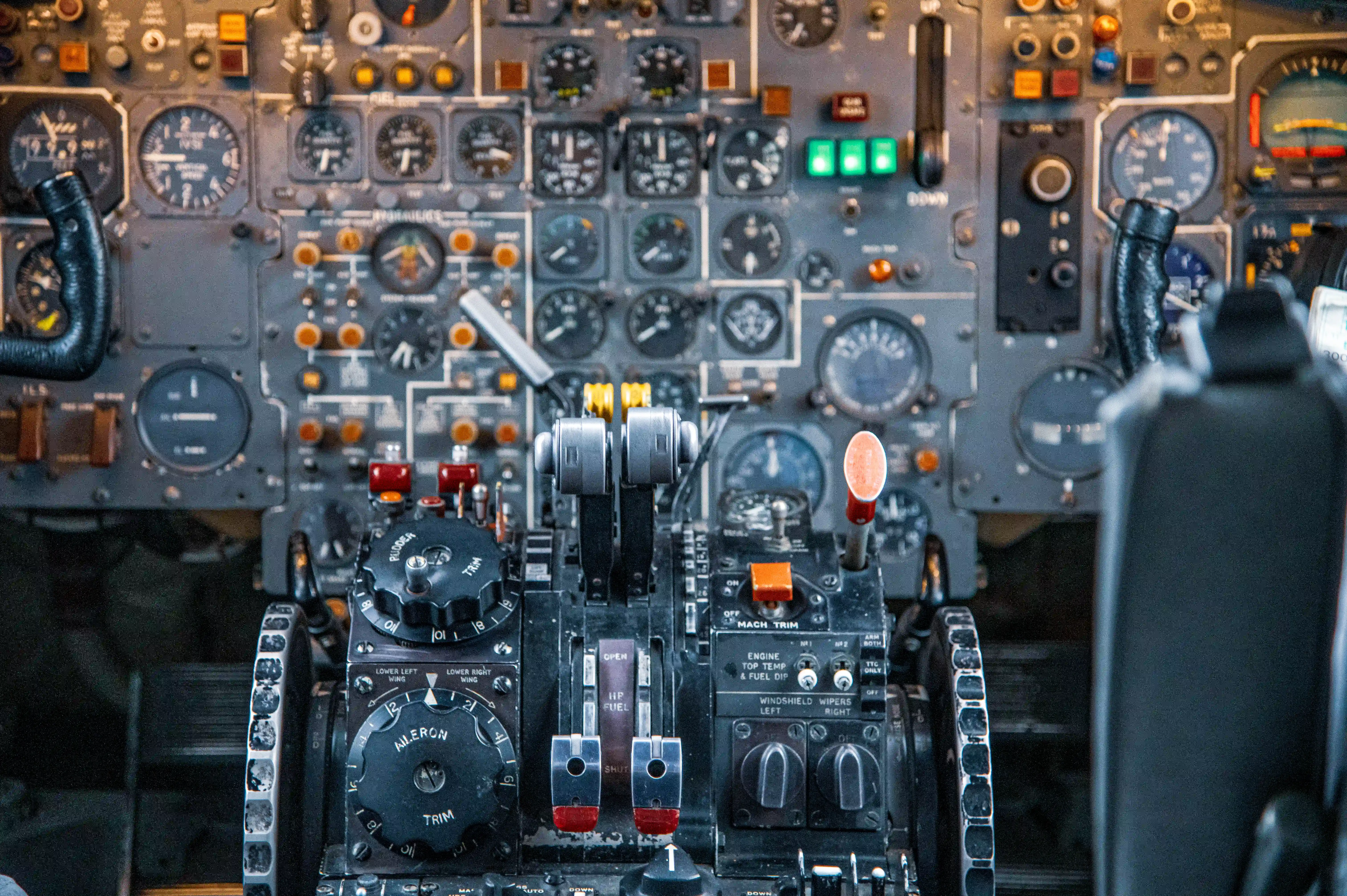 Close-up of an aircraft's cockpit dashboard filled with numerous analogue gauges, dials, switches, and levers, with a focus on the central console containing throttles and trim controls.