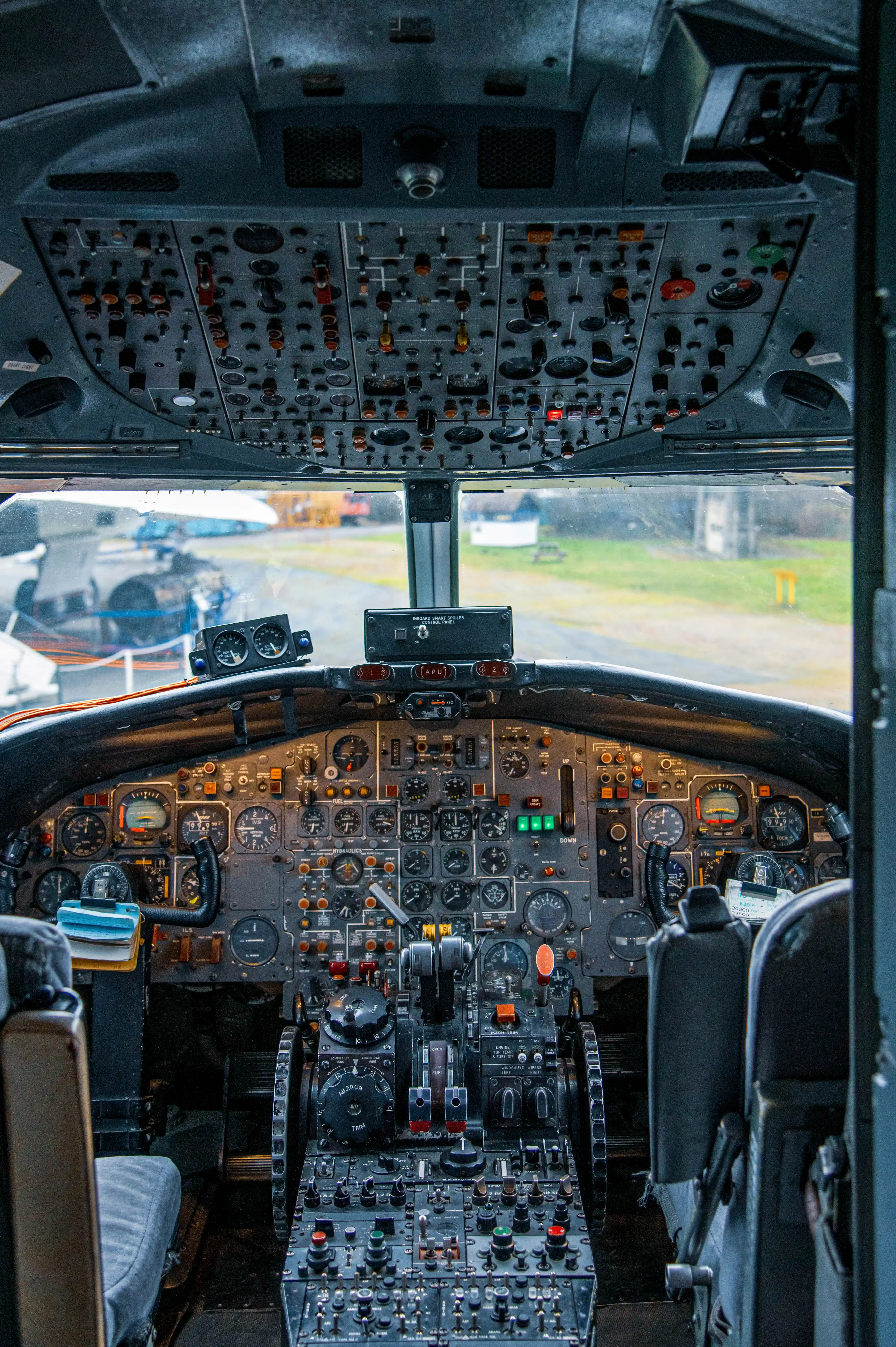 A full view of an aeroplane's cockpit, filled with numerous analogue dials, buttons, and switches. The view looks through the windscreen to an outdoor airfield.