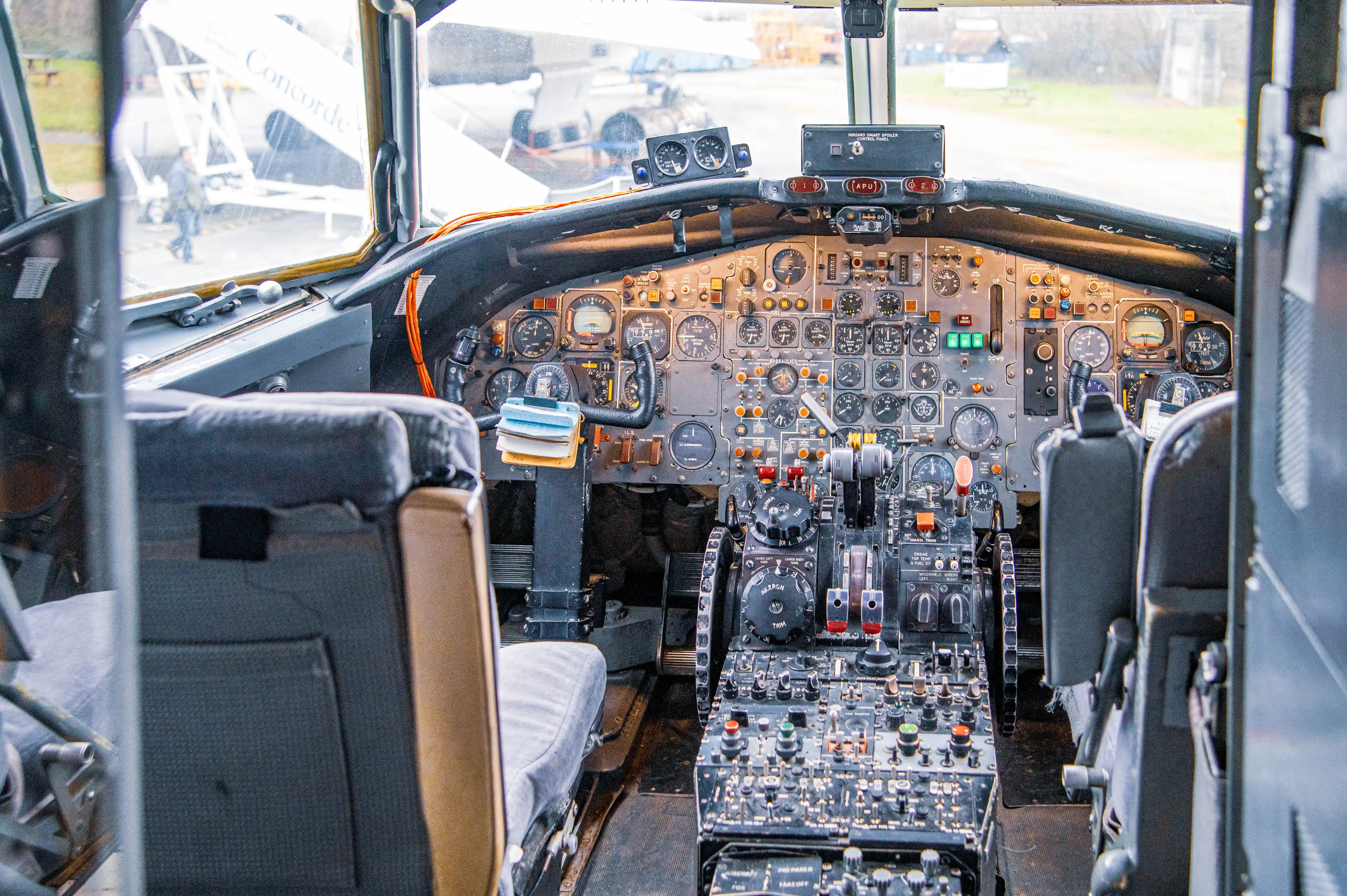 Interior view of the cockpit of a Concorde supersonic aircraft, showing a detailed array of analogue gauges, dials, switches, and levers on the instrument panel. Two pilot seats are visible in the foreground. Through the front windows, another Concorde aircraft is partially visible.