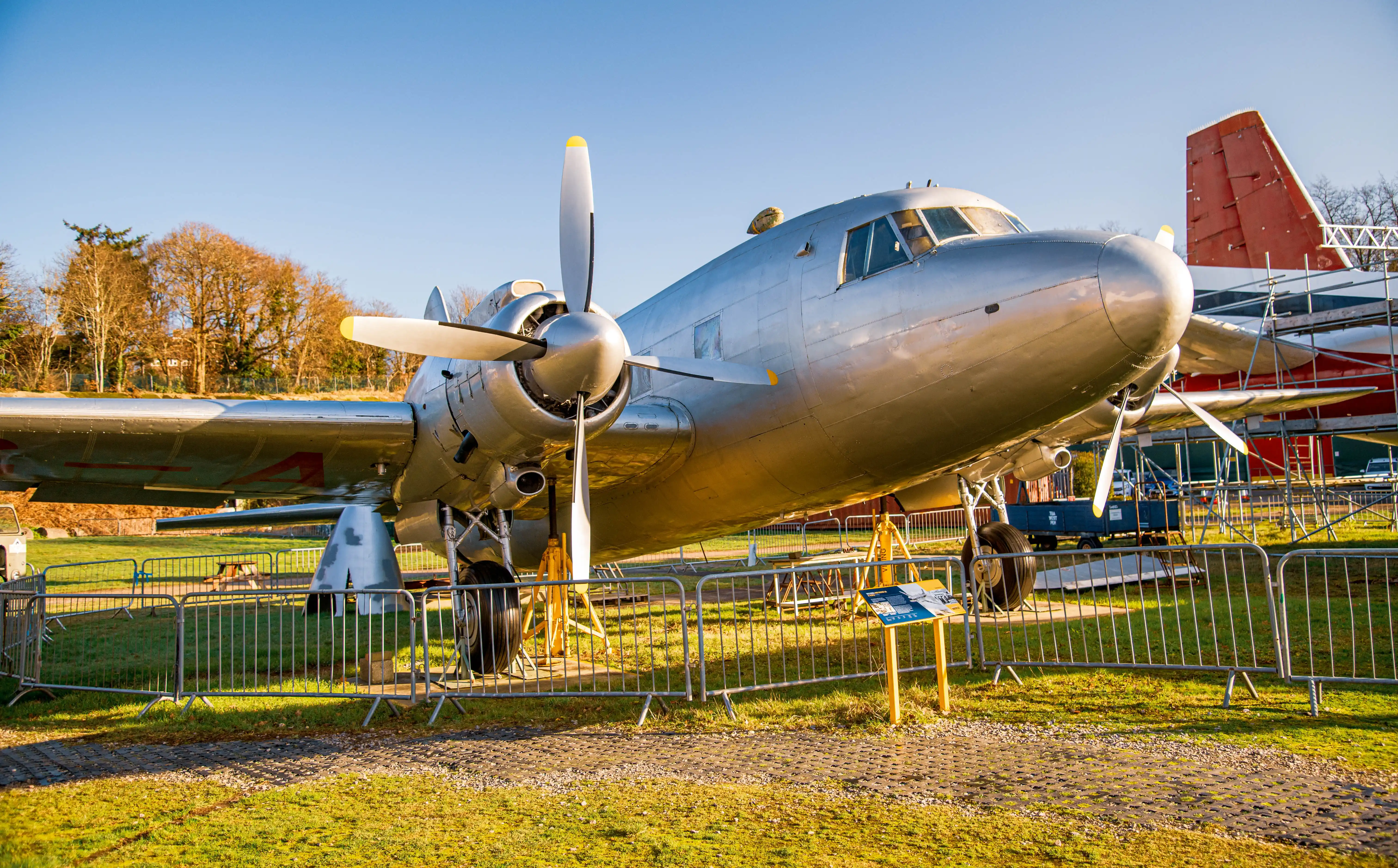 A vintage silver propeller plane with its engine cowling and propellers visible, parked on grass behind a metal fence. Another red-tailed aircraft is behind it, partially covered by scaffolding.