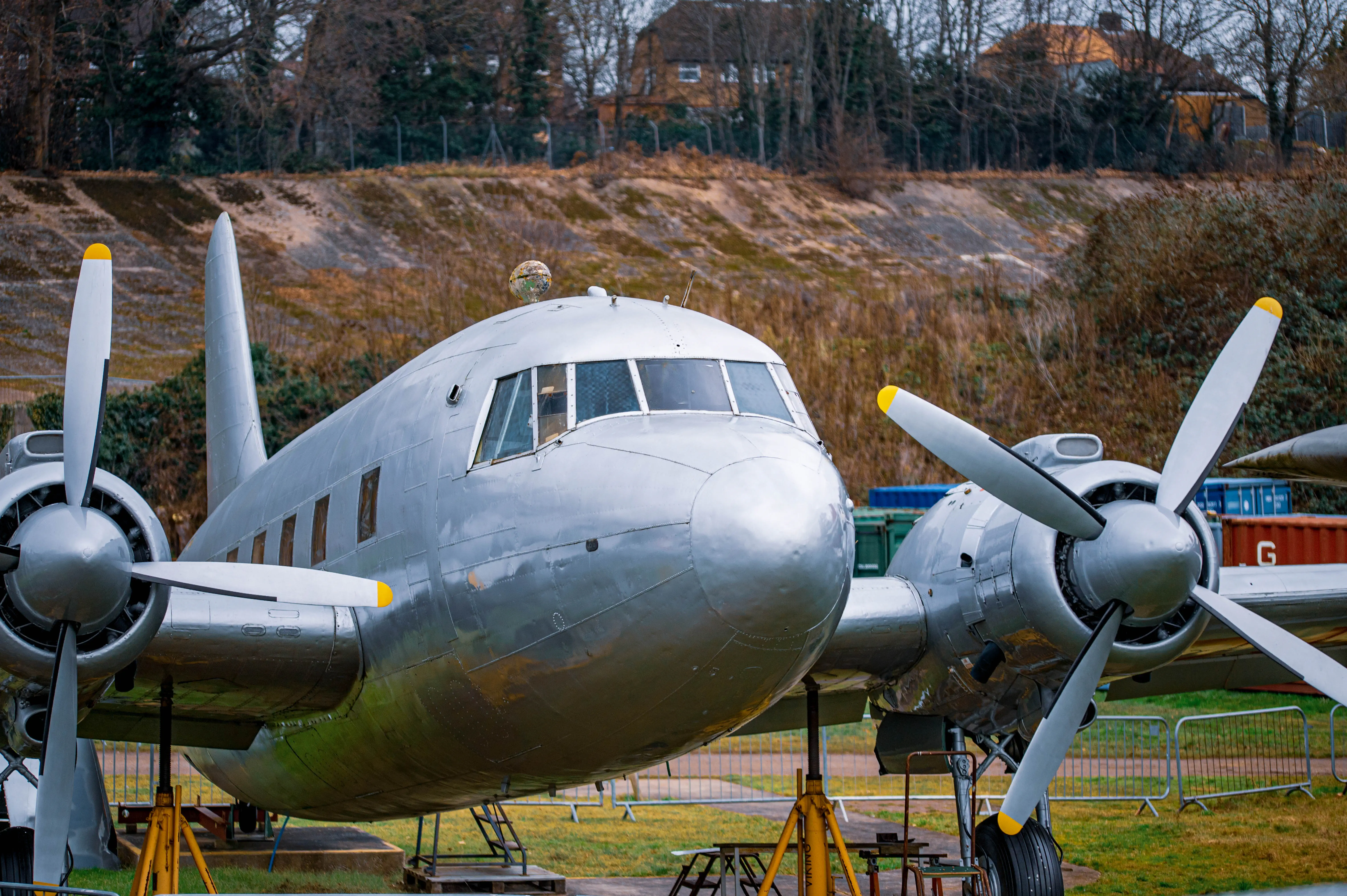 A vintage silver propeller aircraft with two engines and yellow-tipped propellers is parked on a lawn, with buildings and trees in the background.
