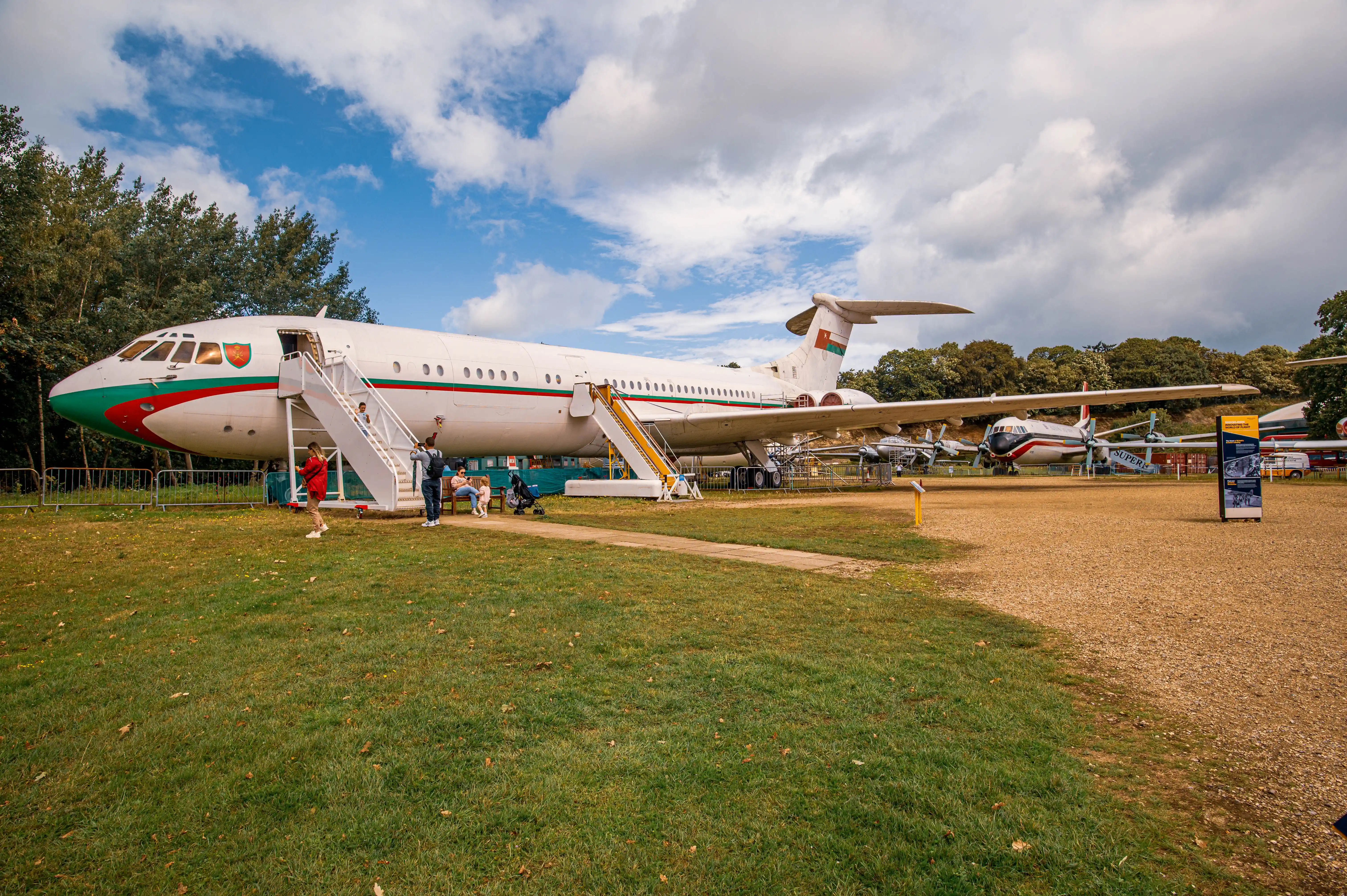 A white passenger jet aircraft with green and red livery is on display outdoors, with visitors standing near its open entrance stairs.