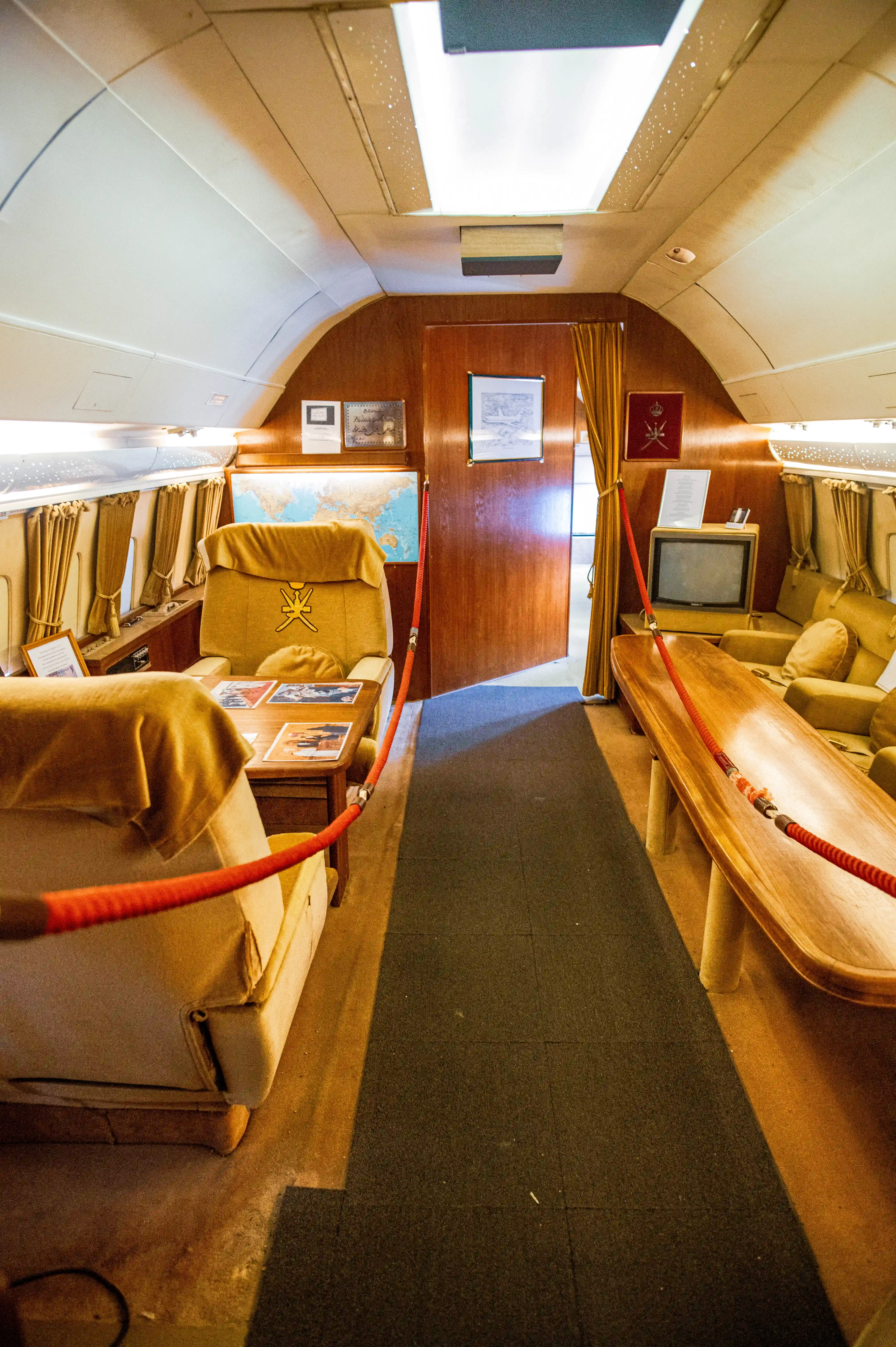 The interior of a private jet cabin, showing two large, comfortable chairs covered in tan fabric. One chair prominently displays a golden crest. The walls are panelled in dark wood, and a world map hangs on the wall.