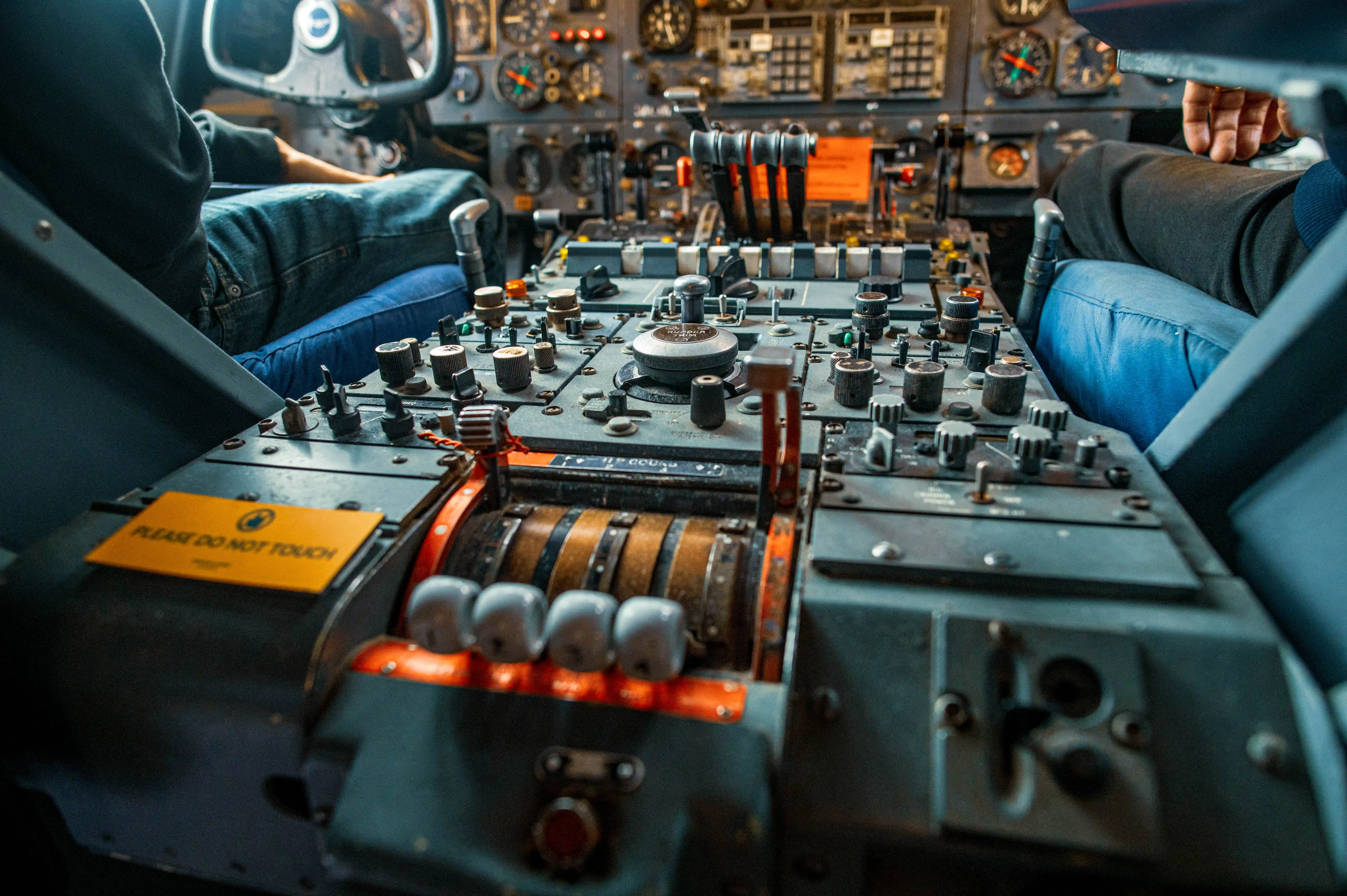 Close-up view of an aircraft cockpit's central control panel with many switches, knobs, and levers, including a throttle quadrant and an orange PLEASE DO NOT TOUCH sign. The legs of two pilots are visible on either side, one wearing blue jeans.